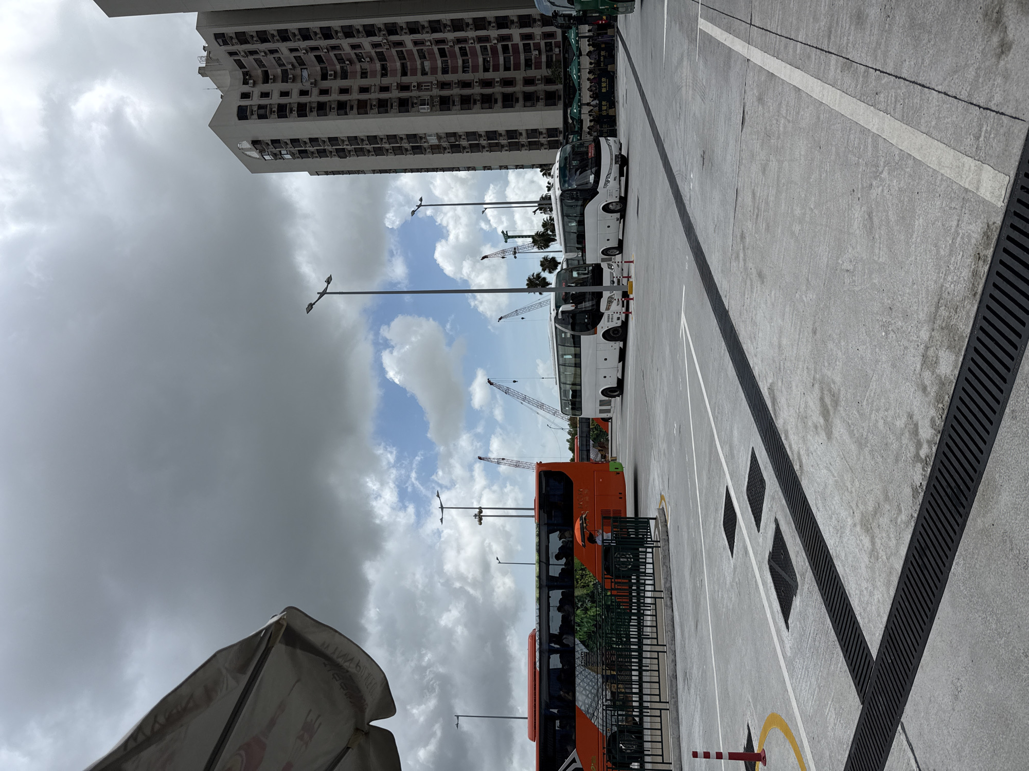 In Macau, China, on a cloudy day, an outdoor bus parking lot is depicted. An orange and green tour bus is parked on the left, with passengers visible inside. Several white tour buses are parked in the lot, and some people are gathered near the entrance of a building on the right. Construction cranes are visible in the background, suggesting ongoing development. The ground is paved with concrete, with visible drainage grates and painted lines. A tall, residential building stands prominently on the right side of the image. A portion of a large umbrella or awning is visible in the upper left corner.
