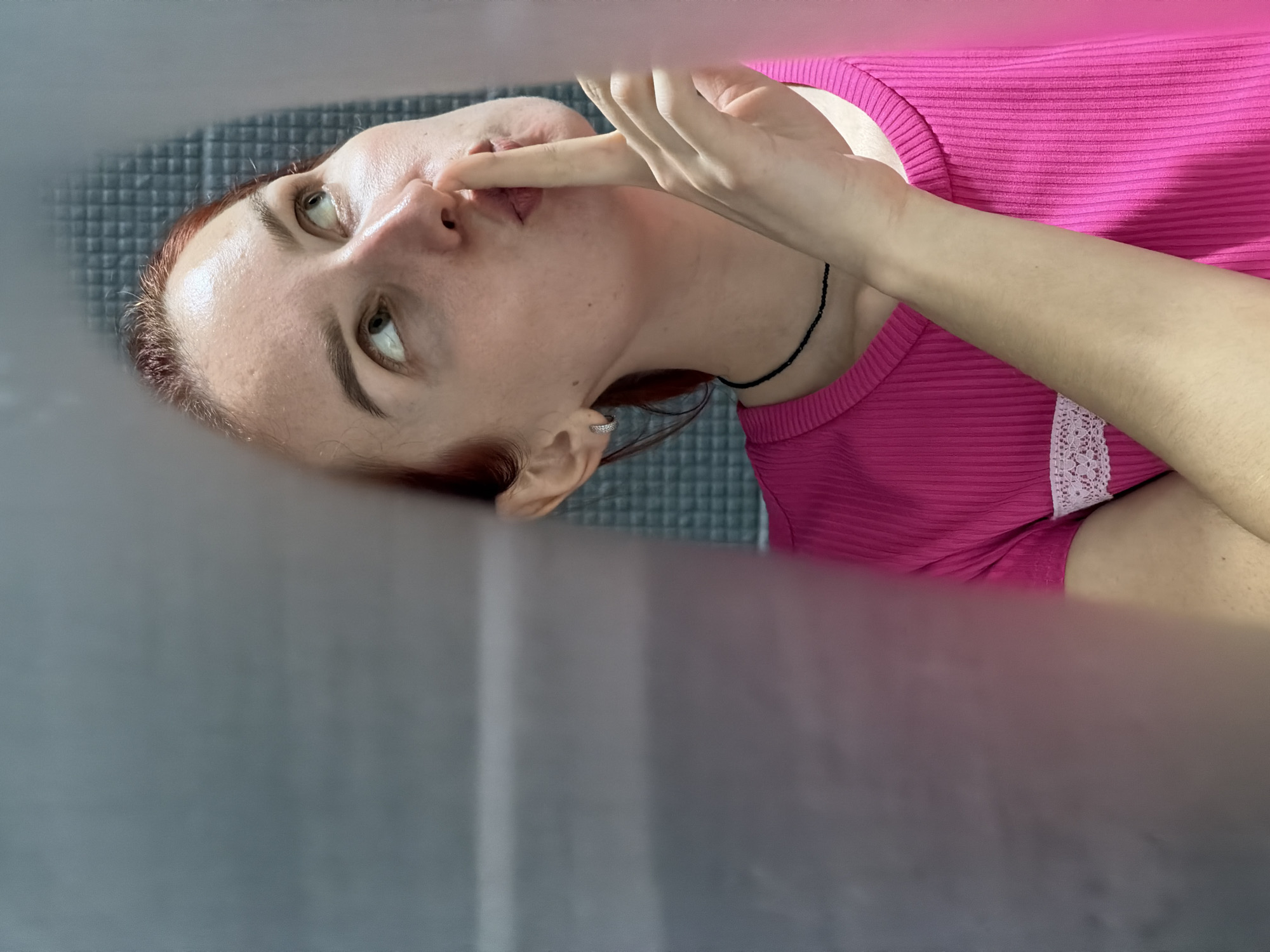 A fair-skinned person with auburn hair and green eyes is lying on a gray, textured surface. The person is wearing a bright pink ribbed shirt with a white lace trim. They are touching their upper lip with their index finger. The background appears to be a blurred gray surface. The location is Chaikovsky, Russia.