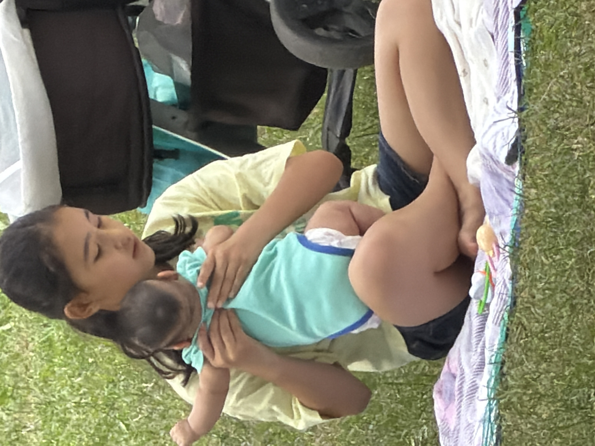 A young girl, appearing to be of pre-teen or early teen age, is sitting on a picnic blanket in a grassy area. She is holding a baby in her arms, with the baby facing away from the viewer and toward the girl. The baby is wearing a turquoise onesie with white trim and a white diaper. The girl is wearing a light yellow t-shirt and dark blue denim shorts. She is looking down at the baby. Behind the girl and baby, there is a stroller with a black bag attached. The ground is covered in green grass, and there are some picnic blankets laid out on the grass. The lighting suggests it is daytime, likely outdoors in a park or a similar setting. The overall scene depicts a tender moment between an older girl and a baby.