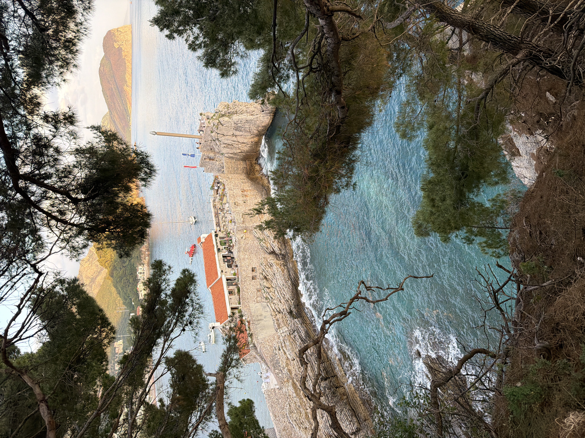 An eye-level photograph captures a view of Petrovac centar, Montenegro. The vantage point appears to be from an elevated position among trees, with the landscape oriented at a skewed angle to the viewer. The azure water is visible, with waves crashing against a stone coast. Adjacent to the water, there is a building with an orange roof, possibly a restaurant. Several boats are docked near the building. In the distance, a mountainous landmass rises out of the water, and the sky is mostly clear. In the upper left and right corners, as well as parts of the foreground, tree branches and leaves obscure portions of the view.