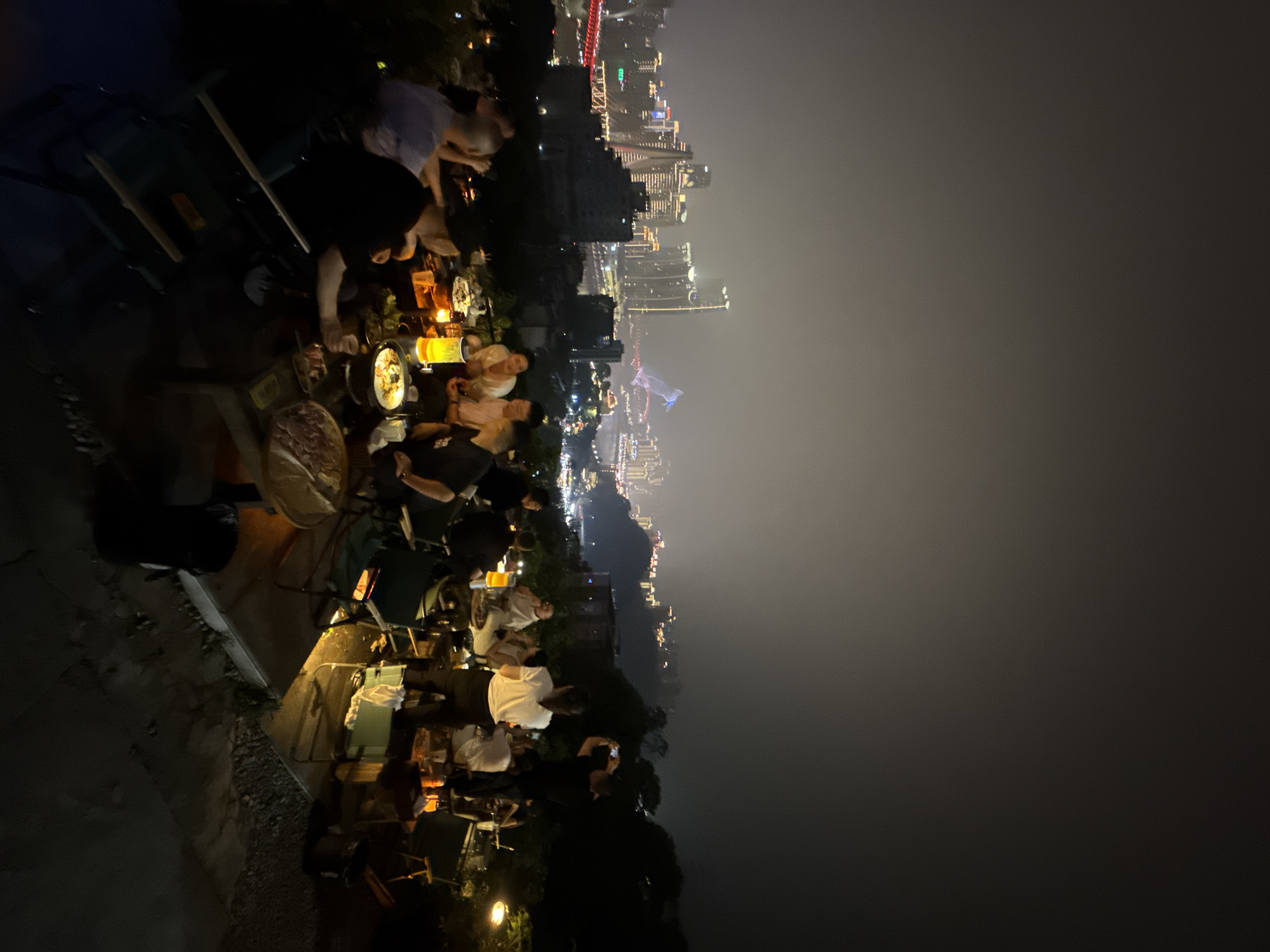 The image depicts an outdoor dining scene at night, likely in the Nan'an District of Chongqing, China. Groups of people sit at tables, eating and socializing. City lights illuminate the backdrop, showcasing a cityscape with bridges and buildings. The tables are set with food, drinks, and lighting. Some people are actively engaged in conversation, while others are using their phones. The foreground shows foliage and the edge of the viewing platform, suggesting a high-altitude or hillside location overlooking the city.