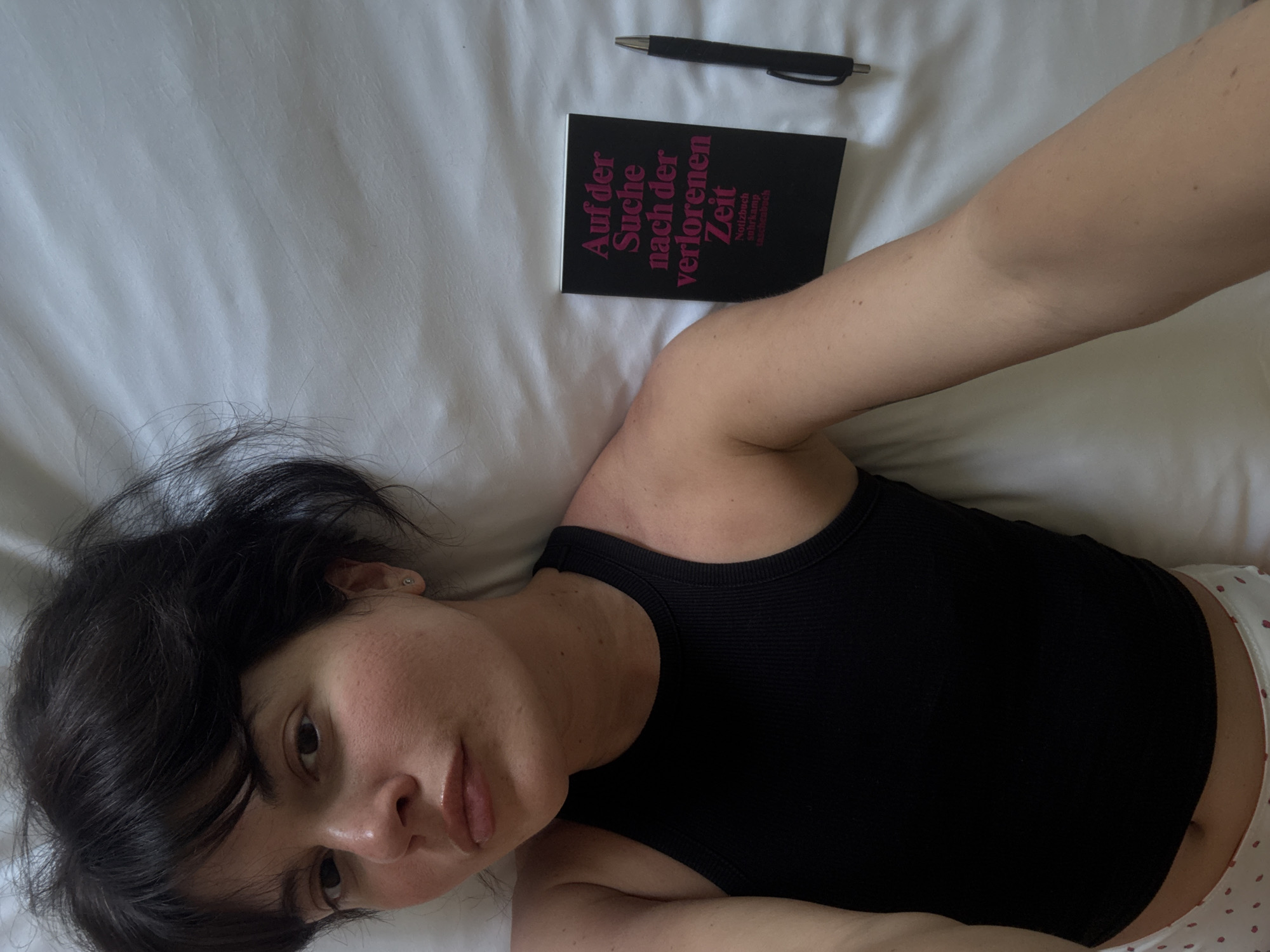 A young woman lies on a white bed, wearing a black tank top and polka-dotted underwear. Next to her is a black book with pink lettering, titled "Auf der Suche nach der verlorenen Zeit," and a black pen. The scene appears to be indoors, possibly a bedroom. There is no indication of the location being in Berlin, Germany.