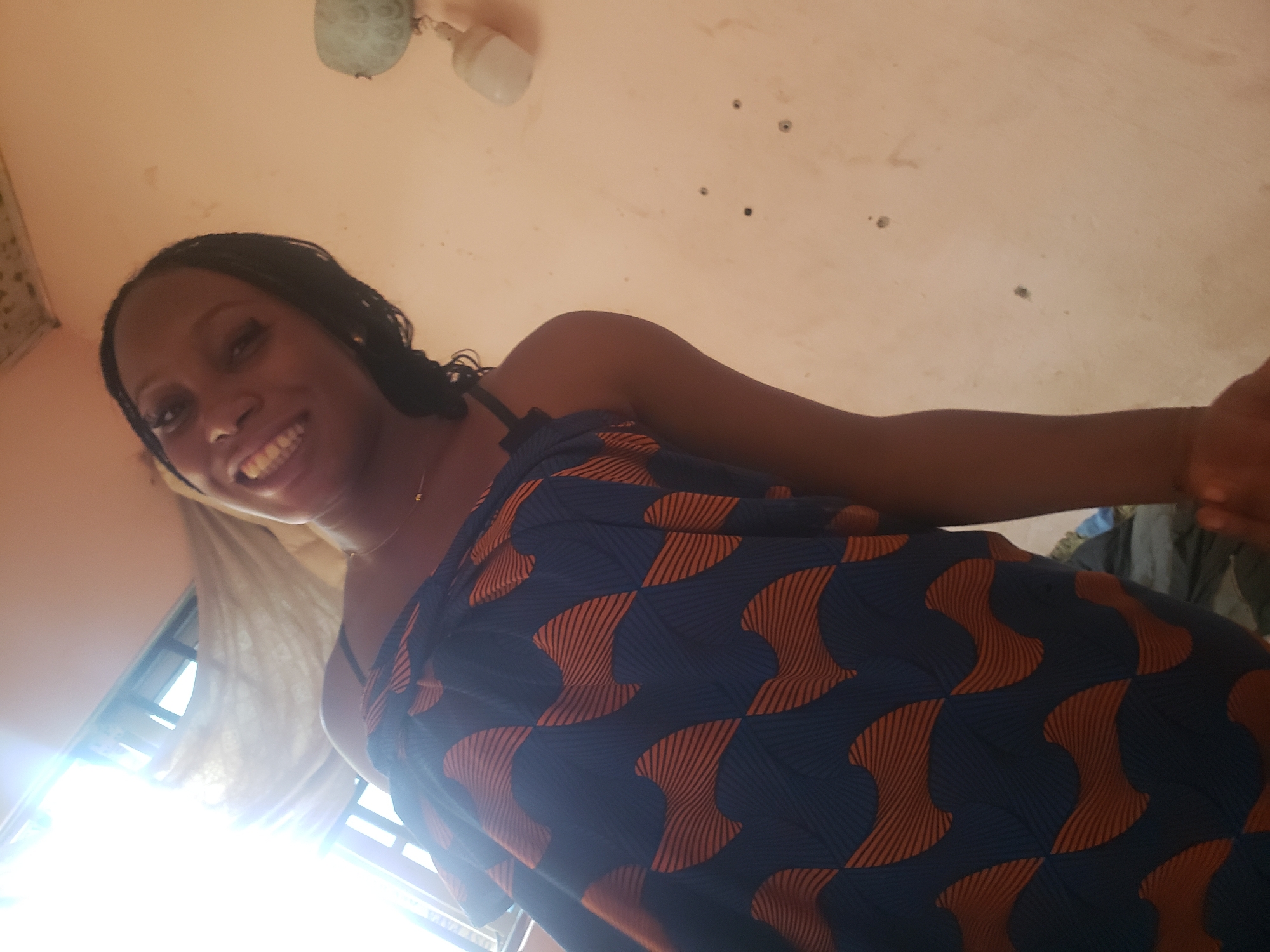 A young woman is the central subject of this upward-angled portrait. She has dark skin, and her hair is styled in dark braids. She is smiling broadly, revealing her teeth, and appears to be looking down towards the camera. She wears a sleeveless garment, likely a wrapper or dress, featuring a striking pattern of deep blue with interlocking orange, wave-like or abstract geometric shapes.
The setting is indoors, with a light-colored, possibly peach or light brown, ceiling forming the background. The ceiling shows some minor blemishes and small dark spots. A ceiling-mounted light fixture with two white bulbs is visible in the upper part of the frame. In the lower left, a strong light source, likely a window, casts a bright glare into the shot, indicating it is daytime. The woman's expression is one of happiness and friendliness. This image depicts an everyday moment in an indoor setting, consistent with the provided location context of Rido, Nigeria. There is no visible text in the image.