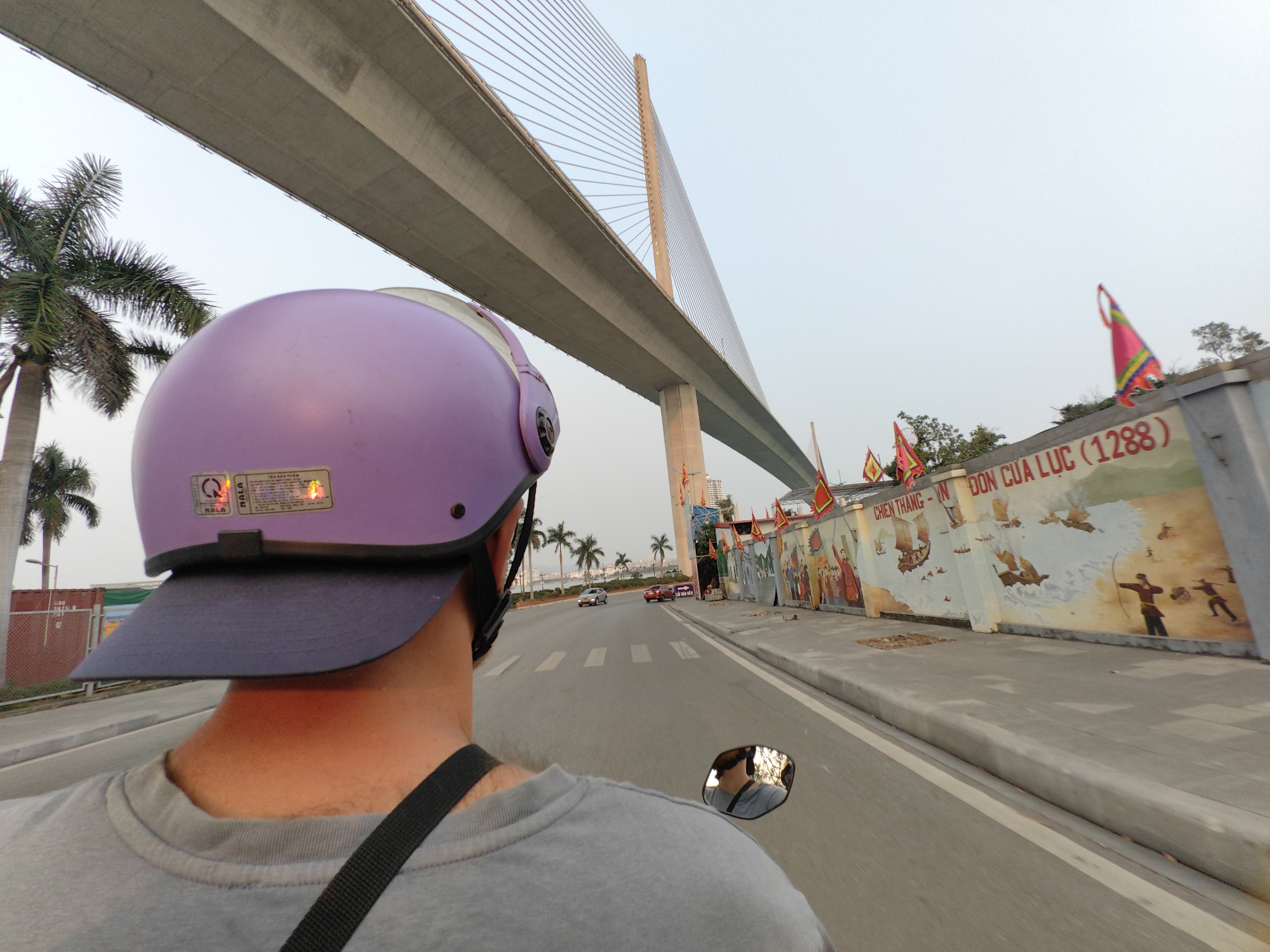 The image shows a person riding a motorbike on a road in Bai Chay Ward, Vietnam. The rider is wearing a purple helmet with a blue brim and a gray shirt. A suspension bridge is above the rider and a palm tree is on the left. On the right side of the road is a concrete wall with painted murals and decorative flags. There are several vehicles driving on the road ahead.