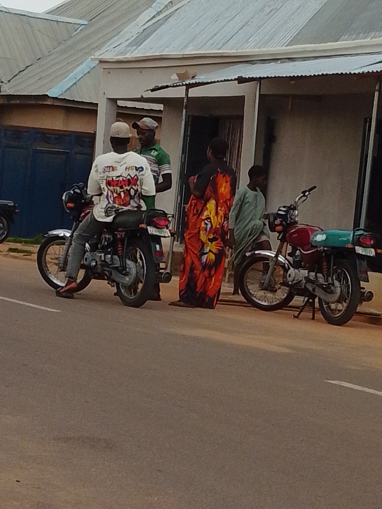 In Tempure, Nigeria, four individuals stand near two motorcycles parked on a paved road. A person wearing a white shirt with a graphic design is seated on a black motorcycle. Next to them, two men in casual attire converse, one with a red cap and the other in a green and white striped shirt. A woman in a brightly colored, patterned dress stands near a red motorcycle, engaging with a person dressed in a light green traditional garment. The background shows a building with a corrugated metal roof and a light-colored facade, suggesting a commercial or residential area. The scene appears to be a snapshot of everyday life in this Nigerian city.