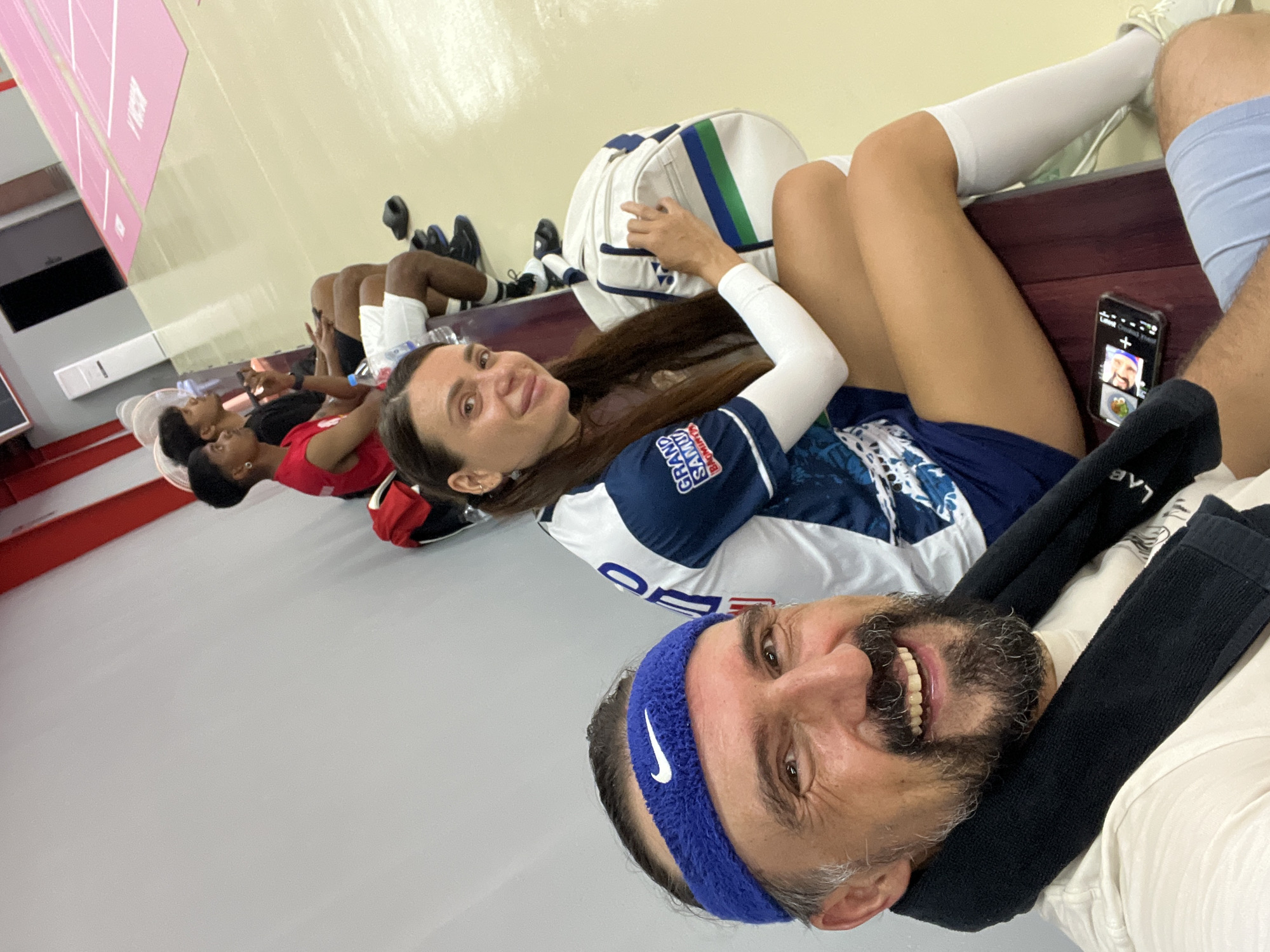 The media file shows a group of people resting on a bench in what appears to be a locker room or a similar indoor facility. In the foreground, a man with a beard and a blue headband is smiling at the camera. He is wearing a white t-shirt and black neck warmer. Behind him, a woman with long dark hair is lying on the bench, also looking towards the camera. She is wearing a blue and white sports jersey and white arm sleeves. A white and green duffel bag is placed beside her.
Further back, several other individuals are seen lying on the bench, some with their eyes closed. One person is wearing a red top, and another is in white shorts and a black t-shirt. Visible on the bench are athletic shoes, including black sneakers and white cleats. A mobile phone is placed on the bench near the woman in the sports jersey, displaying a video call interface with two people.
The scene is indoors with a light-colored wall behind the bench and a grey floor. Red trim is visible along the bottom of the wall on the left side of the frame, suggesting a sports facility. The lighting is bright, indicating daytime. The overall mood appears relaxed and casual, with the individuals resting and possibly taking a break during or after a sporting event or practice.
Text visible on the woman's jersey includes "GRAND SAMUI" and "BLACK MIN." The headband worn by the man has a white Nike logo. A portion of a pink striped surface is visible in the upper left corner, which could be part of a scoreboard or a locker cubby. The location context provided is Ko Samui, Thailand.