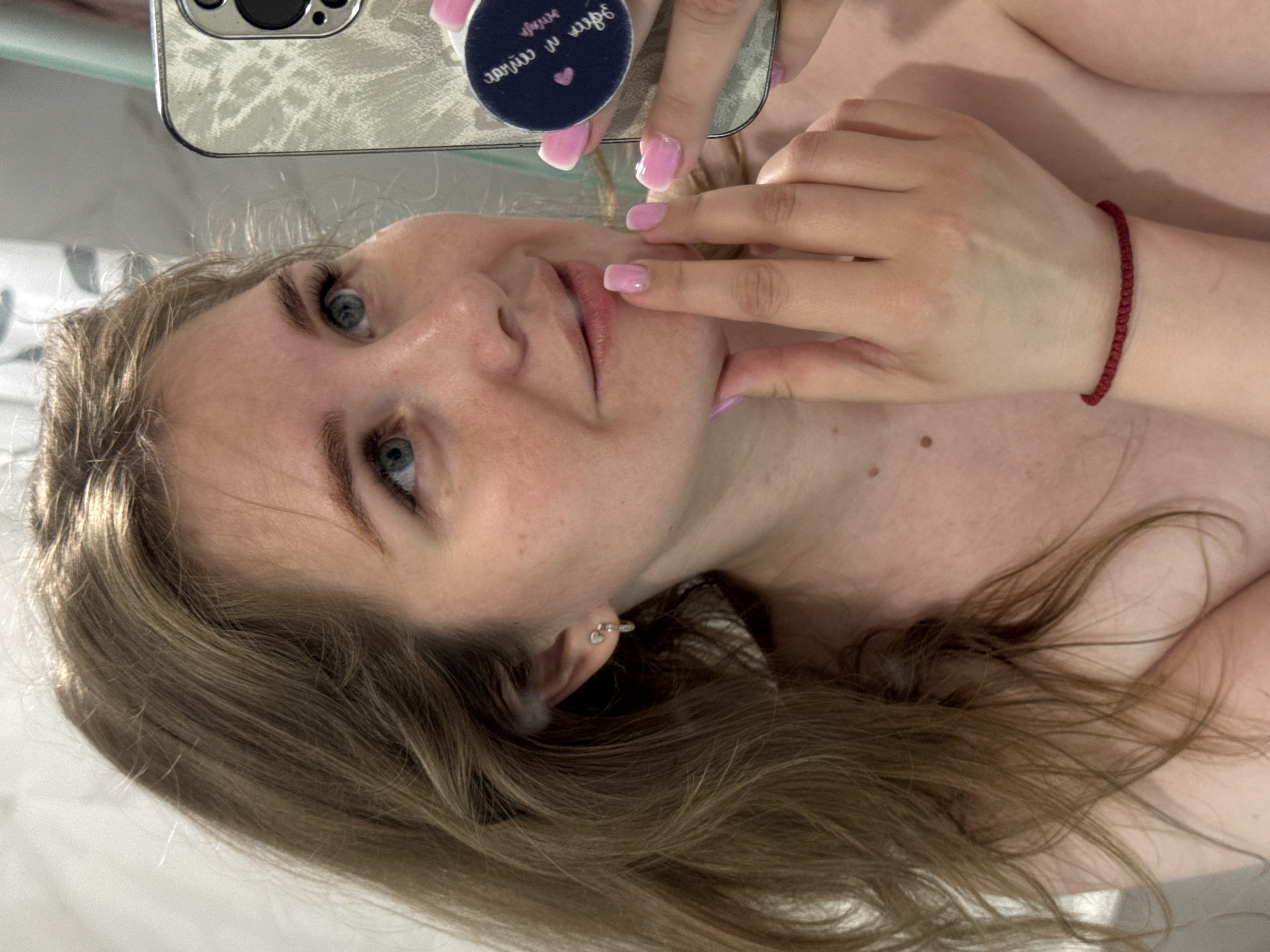 A female individual, appearing to be in her 20s to early 30s, is depicted in a mirror selfie, framed from the chest up. She possesses long, light brown hair with a subtle wave, partially obscuring her left ear, which features a small, silver-toned stud earring with an attached drop element. Her gaze is directed upwards and to the right. Her skin is fair, exhibiting minor freckling on the forehead and cheek, and several small moles on her neck and upper chest. Her lips are naturally pink and slightly parted. Her fingernails are manicured with a pink gradient or French tip style. Her left hand is raised, with fingers touching the area below her lower lip. Her right hand, adorned with a thin red woven bracelet on the wrist, holds a silver-colored smartphone equipped with a textured or patterned case and a dark blue PopSocket. The PopSocket displays mirrored white text, reading "зайти в игру" and "давай" alongside a small pink heart icon. The individual's shoulders and upper chest are bare, indicating minimal or no visible clothing. The background, reflected in the mirror, suggests an indoor environment, possibly a bathroom or bedroom, with a light-colored, subtly textured wall or curtain that includes faint grey or light green patterns. The specific location of this scene in the city of Ферма, Russia, cannot be determined from the visual information present in the image.