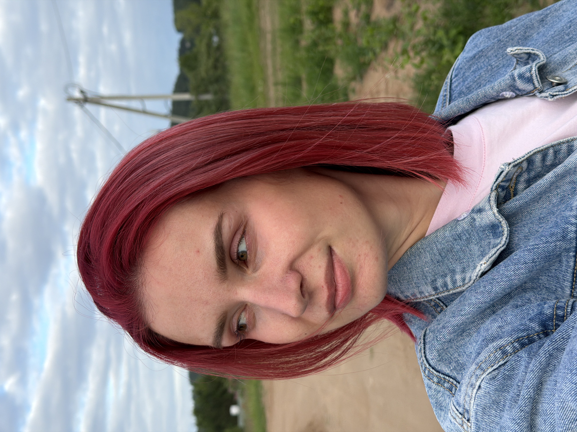 A young woman with vibrant red hair, wearing a light pink t-shirt and a blue denim jacket, is captured in a medium close-up. She is outdoors, with a slightly blurred background consisting of a dirt path, green fields, trees, and a utility pole under a cloudy sky. Her gaze is directed to her left, and she has a neutral expression. The location is identified as the city of Chaikovsky, Russia.