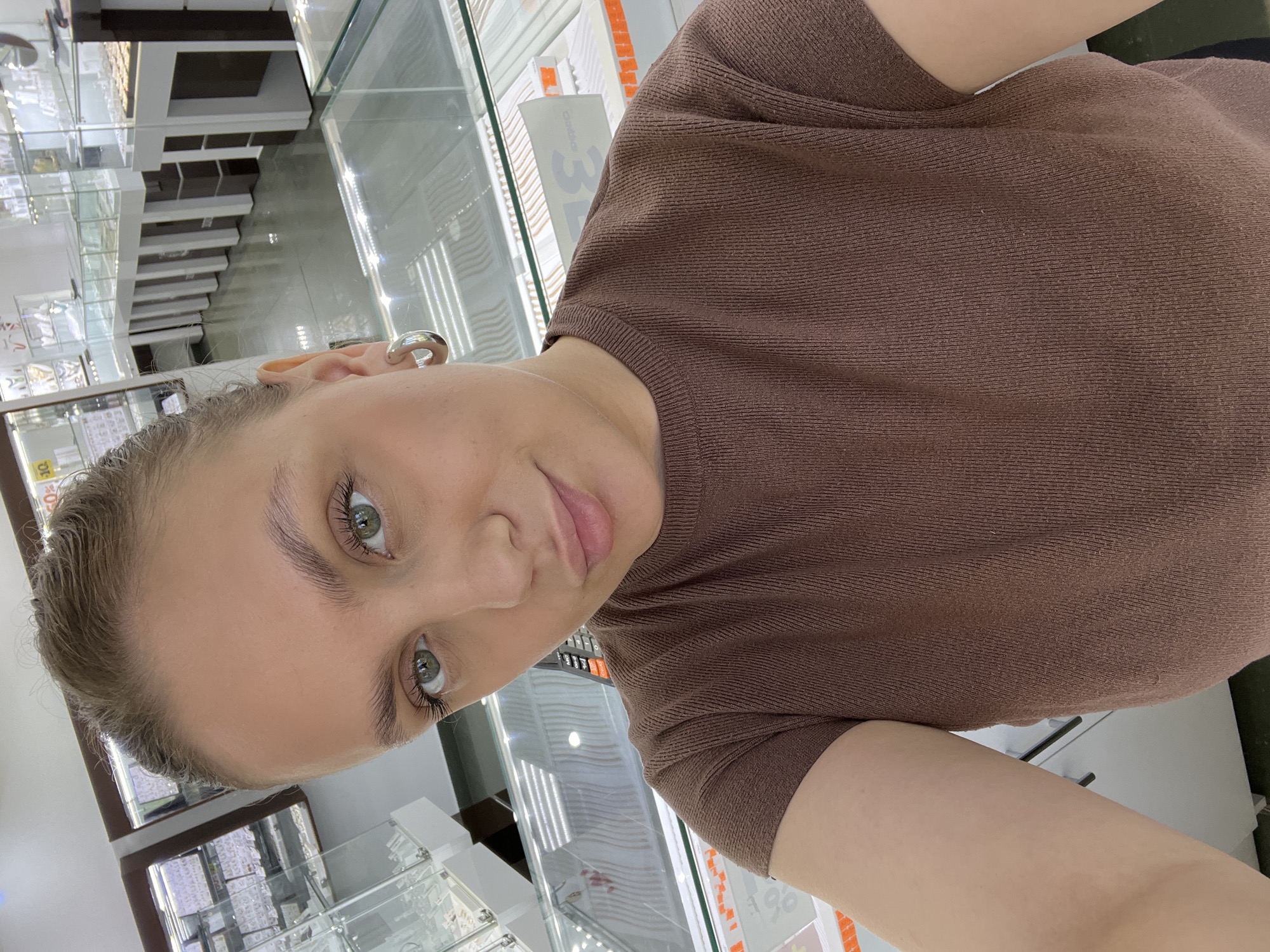 A portrait-oriented photograph, rotated 90 degrees clockwise, depicts a young woman in an indoor retail setting, likely a jewelry store, in Chaikovsky, Russia. She has fair skin, light-colored hair pulled back, and green/hazel eyes, looking directly at the camera with a neutral expression. She is wearing a brown, short-sleeved, ribbed top and a prominent silver-toned hoop earring in her left ear.
The background consists of multiple brightly lit glass display cases filled with various merchandise, which appears to be jewelry or other small items, though specific details are not clear due to distance and reflections. Reflections in the glass show parts of the store's interior architecture and shelving. Visible text on signs in the background includes a prominent number "3" and a red and white sign displaying "90%", indicative of a discount. Another yellow sign shows partial text, possibly related to a percentage. The bright, consistent lighting suggests it is daytime or a well-lit evening within the commercial establishment. No specific activity beyond posing for the photo is discernible.