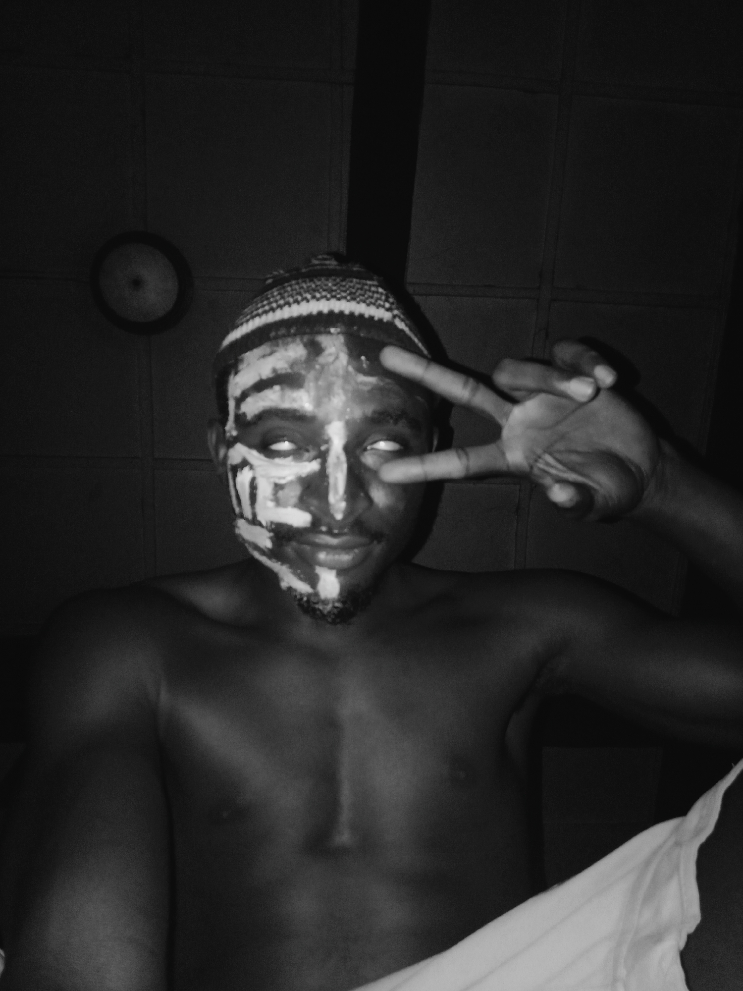 This black and white portrait features a person, likely male, with dark skin, bare-chested, and facing the camera with a slight smile. The central focus is their face, adorned with elaborate white face paint applied in bold, geometric patterns. These patterns include broad strokes across the forehead, outlining the eyes, tracing down the nose, and forming distinct shapes on the cheeks and chin, where a short beard is also visible. The person is wearing a patterned knitted cap or headwear with light and dark zigzags or stripes.
Their eyes are rolled upwards, showing only the whites, adding a striking and intense quality to their expression. The right hand (viewer's left) is raised, forming a peace sign or V-sign with the index and middle fingers.
The setting appears to be indoors with dim lighting, contributing to a mysterious or dramatic atmosphere. A circular object, possibly a clock or a light fixture, is visible on the wall in the dark background to the left. A white piece of fabric or clothing is partially visible in the bottom right corner.
Given the context of Onitsha, Nigeria, the face paint and general aesthetic may suggest cultural, traditional, or artistic expression. There is no visible text in the image.