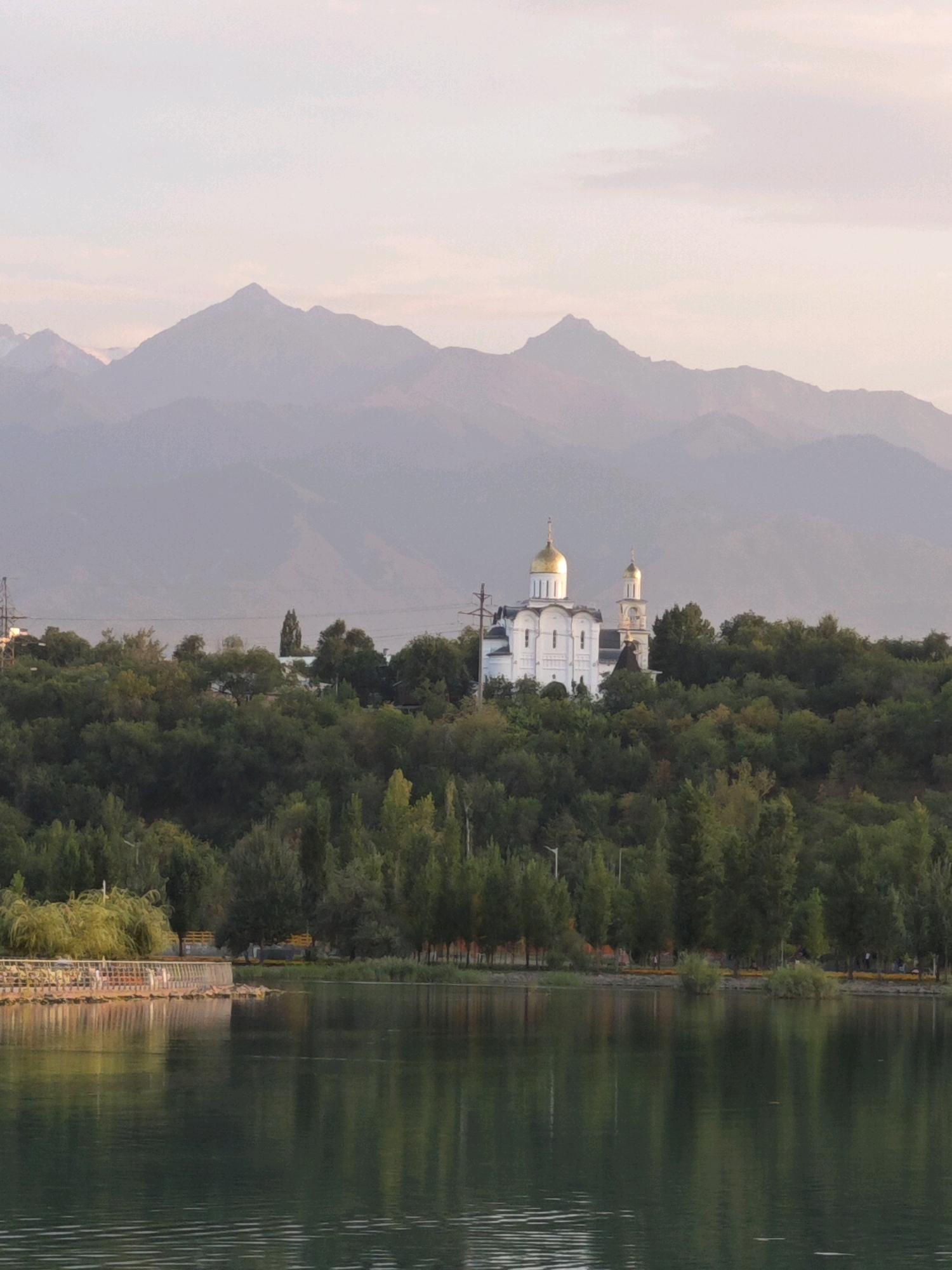 A serene lake reflects the tranquil Almaty skyline, dominated by a prominent white church with golden domes perched on a lush green hill. Towering, mist-shrouded mountains form the distant backdrop, their peaks fading into the soft hues of the sky. The foreground features a gently rippling body of water, with a wooden embankment on the left side of the frame, lined with trees and a few scattered figures enjoying the view. The scene evokes a sense of peace and natural beauty, characteristic of the picturesque landscapes surrounding Almaty, Kazakhstan.
