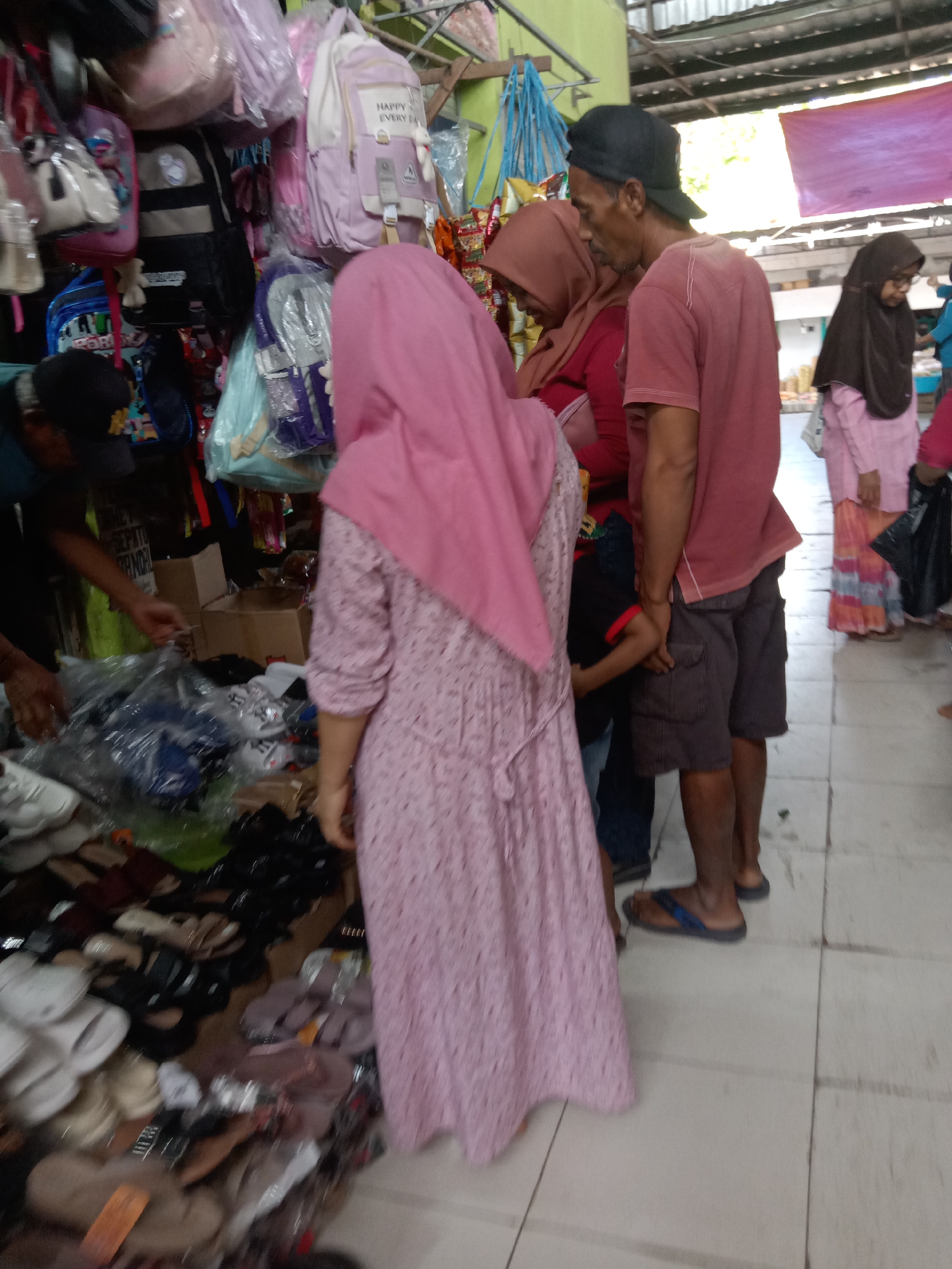 A group of people are gathered in a market stall in Sugihwaras, Indonesia, with an assortment of goods displayed. The stall appears to be selling bags and shoes. In the foreground, a woman in a pink headscarf and dress is looking at shoes laid out on the floor. To her right, a man wearing a maroon t-shirt and grey shorts is standing and looking in the same direction. Partially obscured between them, a child in a black shirt with red trim is visible. Behind the man, another woman in a brown headscarf is also looking at items in the stall. Further back, a woman in a dark hijab and pink dress is standing, holding a white bag. The stall is filled with various bags, including pink and purple backpacks, hanging from the ceiling. A collection of shoes and sandals are arranged on the floor, some in plastic packaging. The environment is an open-air market with a corrugated metal roof visible overhead.