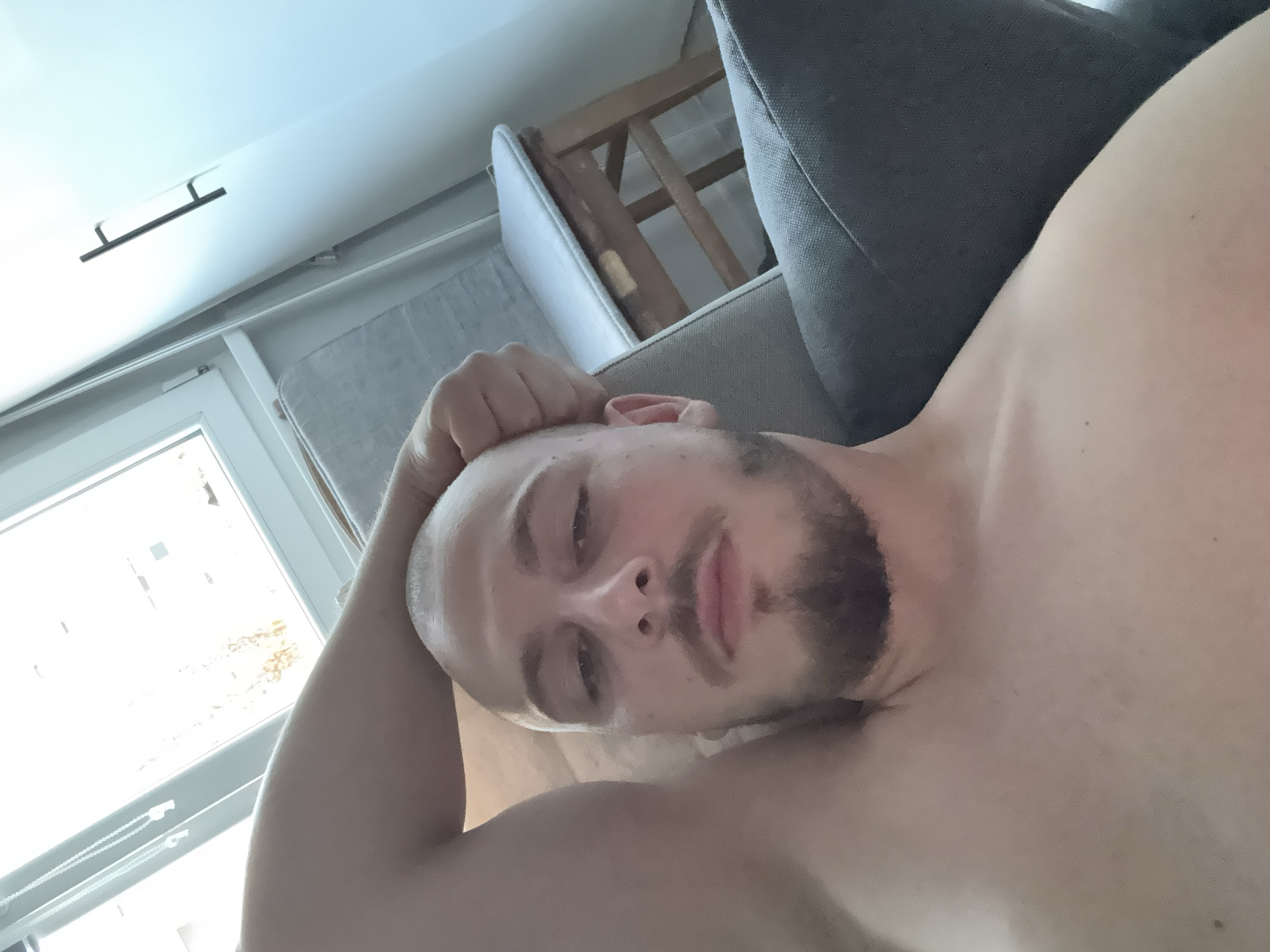 The image is a close-up, first-person perspective selfie taken indoors. The subject, a bald man with a goatee, is lying down, with his arm propped behind his head. He is shirtless, revealing his chest and shoulder. The background shows a window with natural light streaming in, suggesting it might be daytime. Behind him, a part of a chair or sofa is visible, along with a small wooden stool with a white cushion. The overall environment appears to be a residential setting, possibly a bedroom or living room. The image does not provide any specific details that would allow for its location to be identified as Stara Zagora, Bulgaria.
