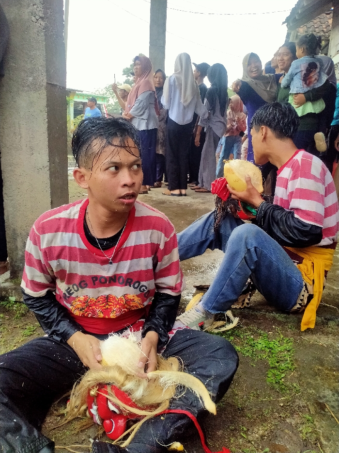 The image depicts an outdoor scene in Tamansari, Indonesia, under an overcast sky, with the ground appearing damp and slightly muddy. In the foreground, two young adult males are seated on the ground. The male on the left, wearing a red and white striped t-shirt with "REOG PONOROGO" text, a black long-sleeved shirt underneath, and black pants, has wet, dark hair and white flecks on his face. He is holding light-colored fibrous material, likely a mask wig, in his hands, positioned over a red, white-faced Reog Ponorogo mask resting on his lap. His gaze is directed towards the right with a wide-eyed, open-mouthed expression. The male on the right, also with wet, dark hair, wears a red and white striped t-shirt, a black long-sleeved shirt underneath, blue jeans, and a yellow cloth sash tied around his waist. He is holding a yellow, half-cut coconut to his mouth, appearing to drink from it. White coconut fragments are scattered on the ground around him. Behind these two individuals, a crowd of approximately 10-15 observers, primarily women wearing various colored headscarves and children, stands in the mid-ground. One woman on the far right holds a small child. Concrete pillars are visible on the left and in the mid-background, with a building featuring a dark roof and a green-trimmed building in the far background. Small patches of green vegetation are interspersed on the damp ground.