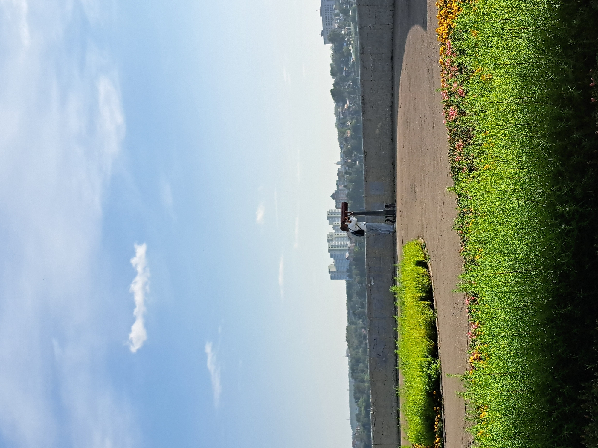 The image captures a daytime scene in a park area, likely along the Angara River in Irkutsk, Russia. In the foreground, a bed of vibrant green foliage and some blooming flowers, in shades of yellow and pink, lines a curved paved walkway. A concrete barrier separates the walkway from a viewpoint overlooking a distant cityscape. In the middle ground, a person, dressed in light-colored pants and a white shirt, stands behind a large, coin-operated telescope, looking through it. The person is facing away from the camera, towards the distant buildings. The background reveals a panorama of a city with various buildings, including several high-rise structures, nestled among trees. The sky above is a clear blue with a few wispy white clouds. The overall impression is one of a pleasant, open public space with recreational facilities.