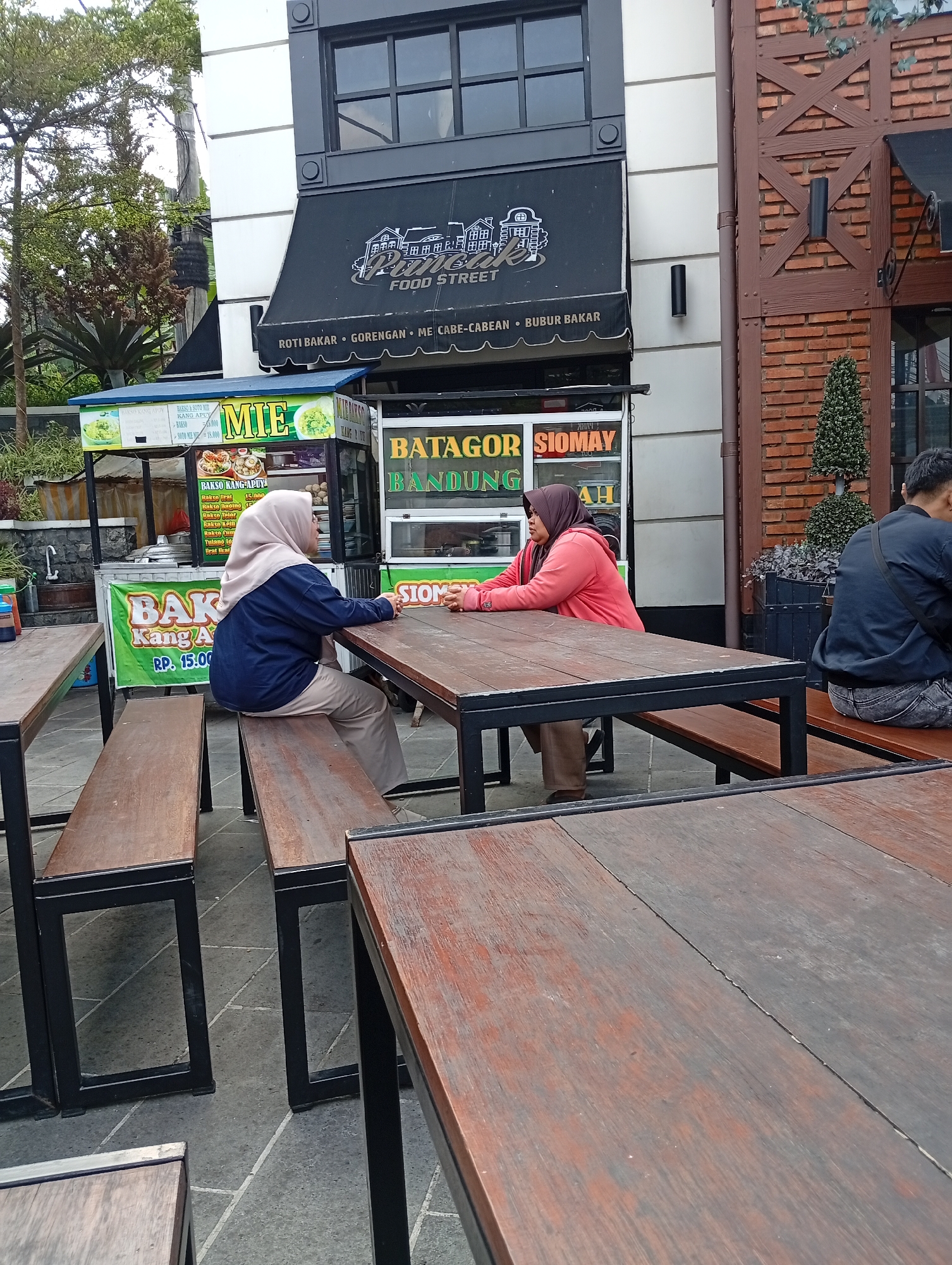 Two women are seated at an outdoor wooden table in front of food stalls in Cimacan, Indonesia. The woman on the left wears a blue jacket and beige hijab and sits on a wooden bench. The woman on the right wears a pink hoodie and brown hijab and also sits on a bench. Behind them, food stalls display menus for "Mie" (noodles), "Batagor," and "Siomay." The stalls have signage with prices and food item names. The area appears to be a marketplace or food court. In the background, there's a building with a black awning and windows, and some greenery.