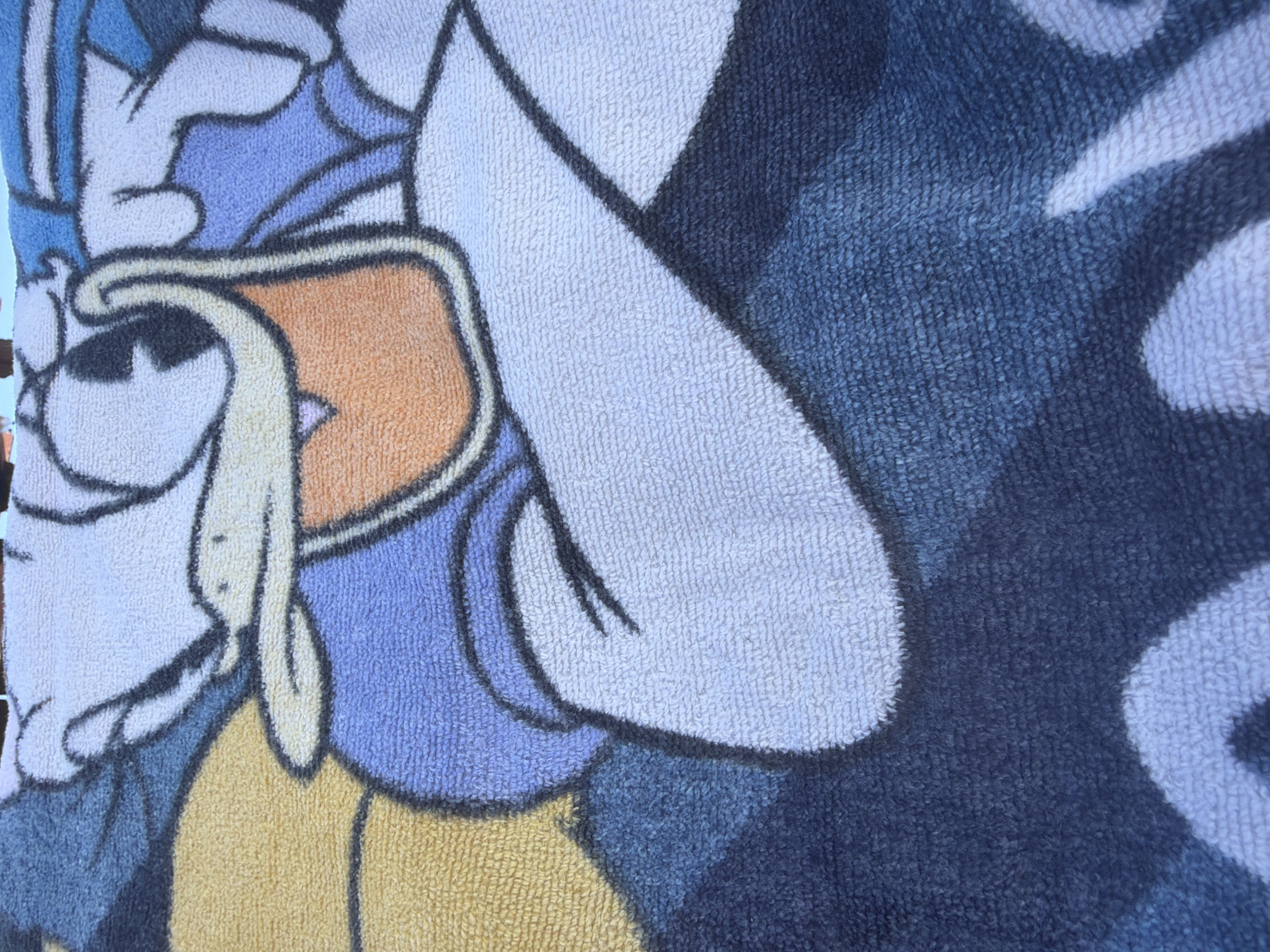 A close-up, high-angle shot of a towel with a cartoon character printed on it is shown. The towel is made of terry cloth and features the character Donald Duck. The towel is folded and rests on a dark blue surface, which is likely a floor or bed. The character is depicted from the chest up, with his distinctive sailor suit and large beak prominently displayed. The colors are vibrant, with blues, white, yellow, and orange used to create the image. The texture of the terry cloth is visible, giving the image a soft and tactile feel. The scene is likely domestic, possibly in a home or hotel room. No specific location in Sharm El Sheikh, Egypt, can be identified from the image.