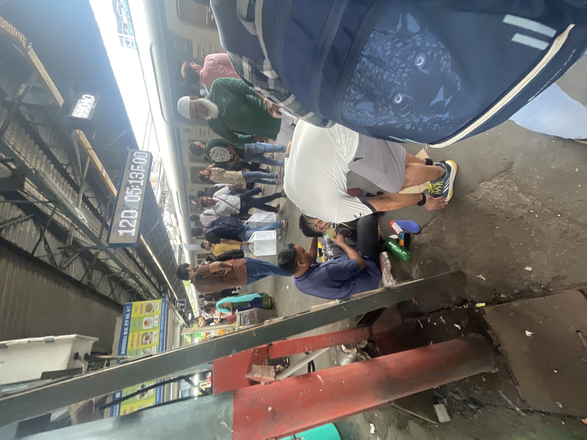The image captures a bustling scene at a train station in Mumbai, India, likely Borivali railway station given the sign visible in the background. In the foreground, a man in a blue shirt is intently focused on something in his hands, possibly working on a small object or performing a task that requires close attention. He is seated on the ground, surrounded by various items including a plastic bottle, a small white box, and what appears to be a pair of scissors. Nearby, another person in a white t-shirt and grey shorts is bending down, their attention also seemingly directed towards the ground.
The environment is a typical Indian railway platform, characterized by the presence of a train on the tracks, a covered waiting area with a corrugated roof, and overhead signage displaying station information and train schedules, including digital clocks showing "12D 05:13:00" and "59040." People are milling about, some carrying bags and others engaged in conversations, creating a sense of movement and activity. The ground is concrete and appears somewhat worn, with scattered debris suggesting a busy public space. The overall impression is one of everyday life unfolding at a busy transportation hub in Mumbai.