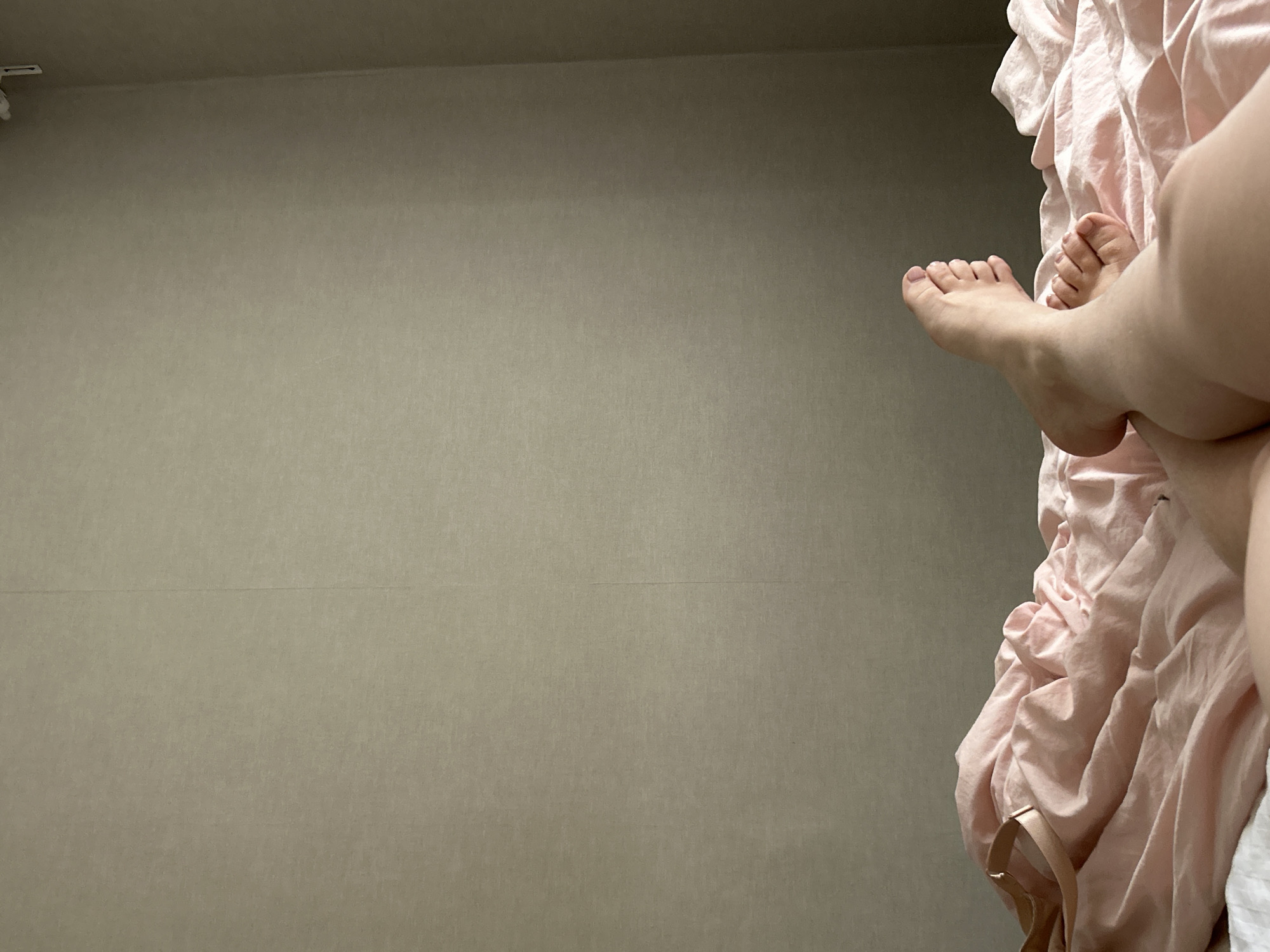 The image captures an indoor scene, likely a bedroom, from a low angle, focusing on a person's lower legs and feet against a plain wall. On the right side of the frame, two feet and parts of the lower legs are visible, suggesting the person is resting or relaxing on a bed. One foot is more prominent, with toes clearly visible, while the other is partially obscured behind it.
The person's legs are partially covered by or resting upon a large amount of soft, light pink, wrinkled fabric, which appears to be a bedsheet or blanket. Tucked into this fabric near the bottom right, a narrow, light-colored strap, possibly from an undergarment, is visible. The background is a textured, uniformly colored wall, appearing in shades of light greyish-brown. In the top-left corner, a small, white, L-shaped fixture, possibly a sensor or bracket, is attached near the ceiling line.
The lighting is soft and diffused, characteristic of indoor ambient light, creating a calm and relaxed atmosphere. There are no direct indications of the time of day or outdoor weather conditions. No visible text is present in the image. The setting is in Chaikovsky, Russia.
