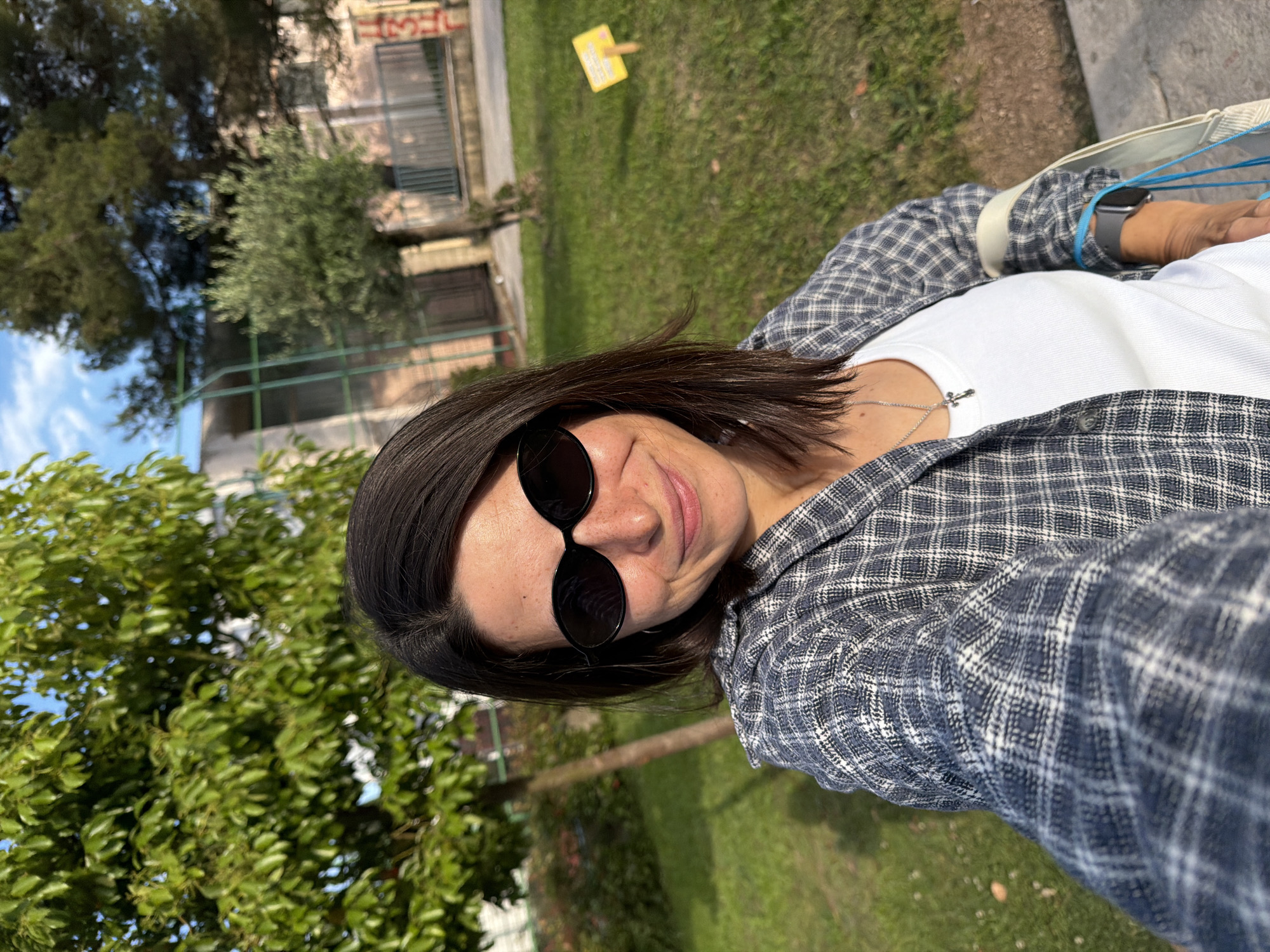 A medium shot of a woman in a public area in Budva, Montenegro. The woman is wearing dark sunglasses, a white shirt, and a plaid jacket. She also has a silver necklace with a cross pendant. She is facing the camera and appears to be taking a selfie. The background includes a grassy area, trees, and a building in the distance.
