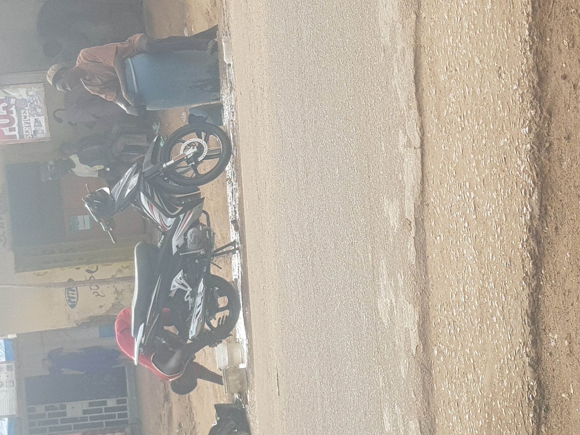 The scene is an exterior shot of a street in Tukur Tukur, Nigeria. A black and silver motorcycle is parked on the right side of the street. A man in a red shirt is crouched down near the motorcycle. Nearby is another man in a tan shirt and blue clothing is visible near a building. There is a group of people in the background near a building with a sign that says "SERVICE". The street is paved.