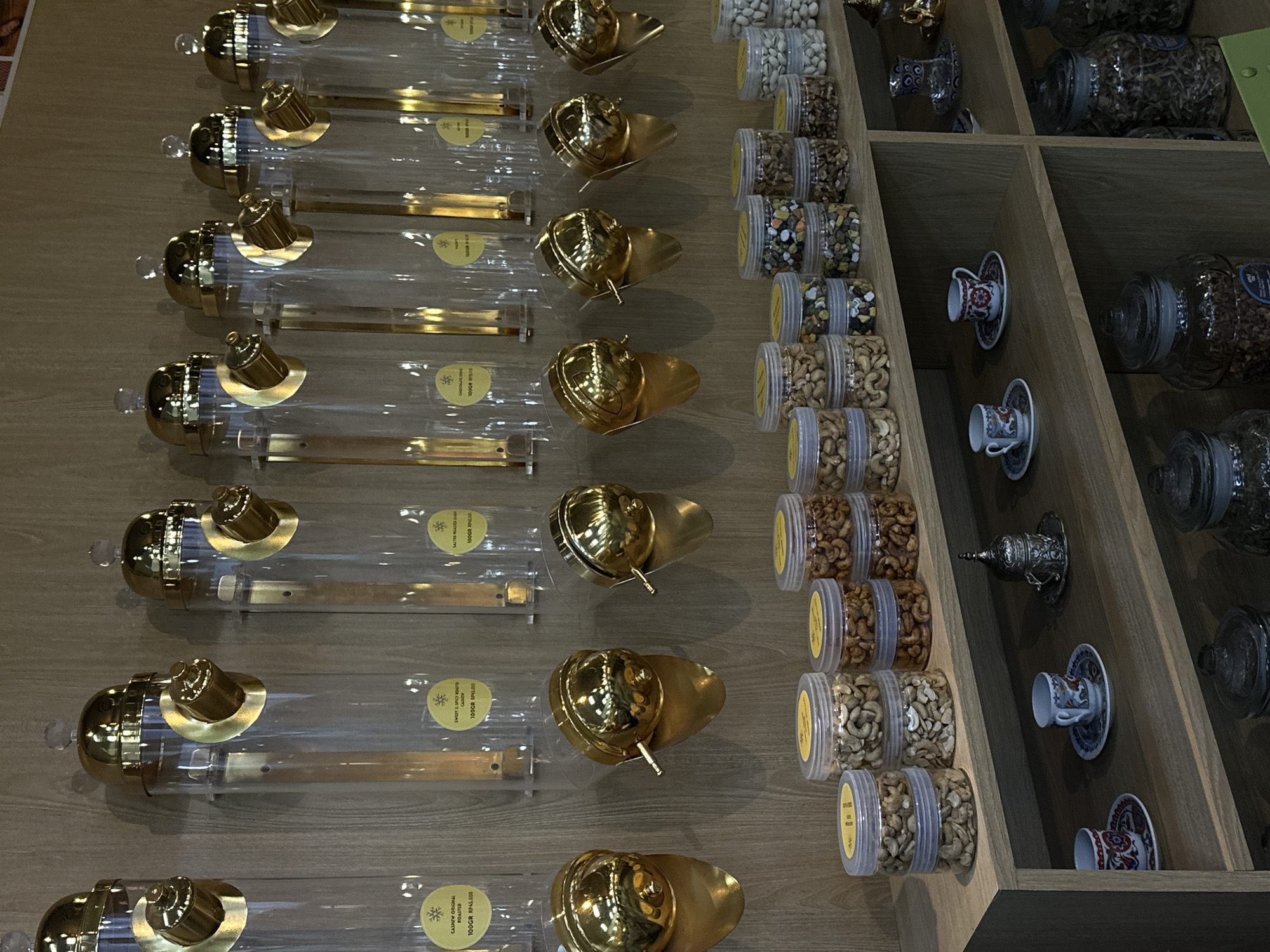 The image shows a display of gourmet snacks and coffee items in a store, possibly a specialty food shop or gift shop. The items are arranged on a wooden shelf unit. On the left are cylindrical clear containers with golden lids and dispensing mechanisms. They are filled with what appears to be saffron or other expensive spices. Each container has a small yellow label with text that is partially obscured, but includes the price "100GR RP45.000." Next to these are rows of clear plastic jars filled with various types of nuts or seeds, likely for sale. In a separate section of the shelf are small, decorative espresso cups and saucers, along with glass jars filled with coffee beans or tea leaves. The scene is orderly and well-lit, suggesting a curated retail environment. While the image is not a traditional crime scene, it documents the inventory and layout of a retail space, potentially useful for investigative or auditing purposes. North Kuta, Indonesia, is mentioned as the location.