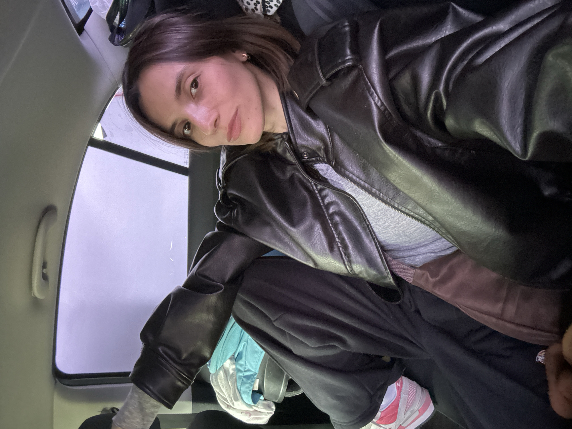 A young woman with brown hair is seated in the back of a vehicle. She's wearing a gray t-shirt and a black leather jacket. She has on black pants, with a glimpse of a brown garment underneath. She's wearing a white sneaker with red accents. The interior of the vehicle is visible, including the window, a white grab handle, and various items like clothes and headphones. No definitive landmarks indicate the location is Moscow, Russia.
