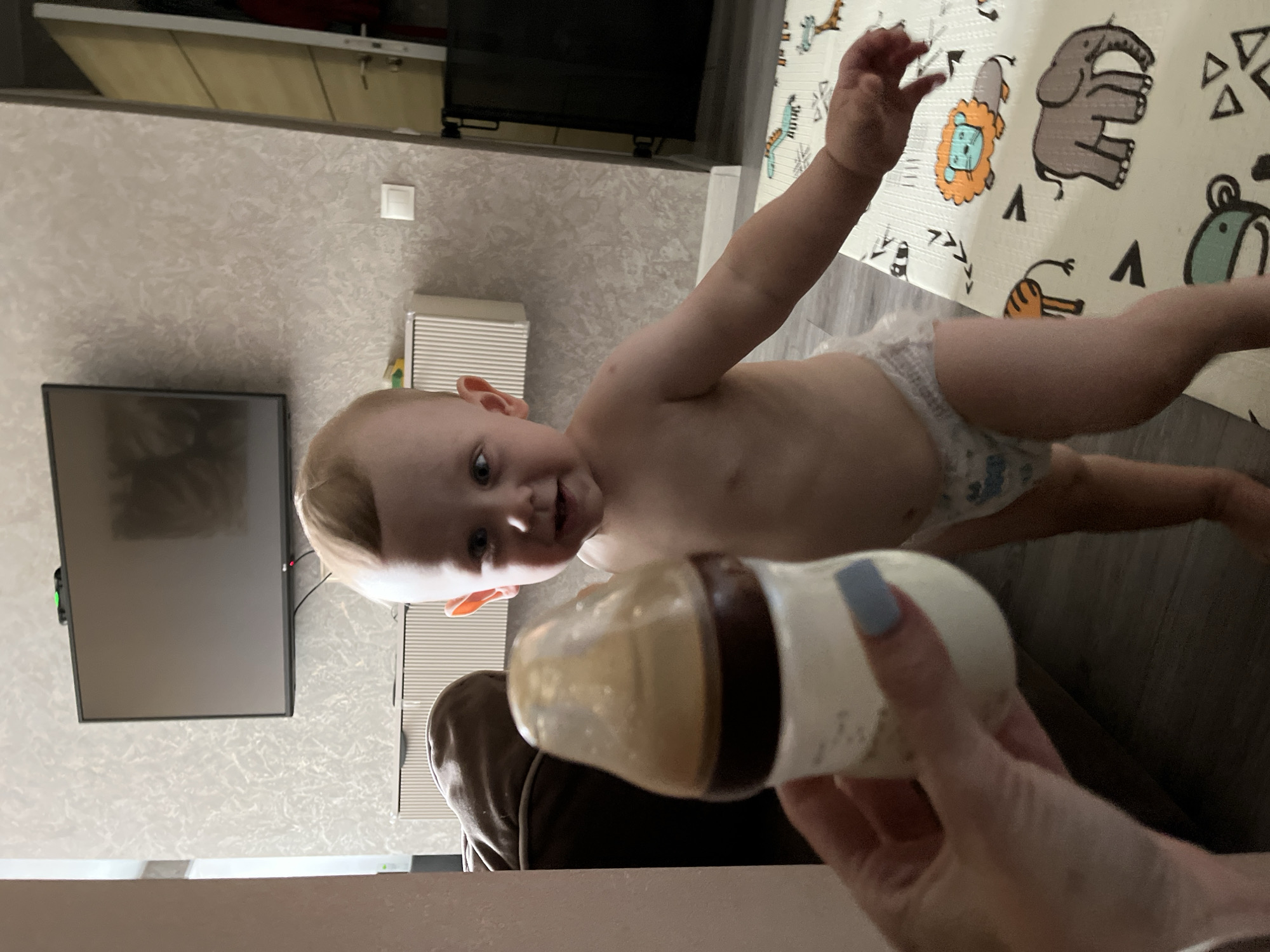 In an indoor setting, likely a residential apartment, a toddler stands wearing a diaper. The toddler is in the middle of the frame, facing the camera with a slight smile. In the foreground, a hand, presumably belonging to an adult, holds a baby bottle filled with a light brown liquid. The background features a wall with textured wallpaper, a flat-screen television mounted on the wall, and a cabinet. To the right, a partially visible doorway leads to another room. A colorful playmat with animal illustrations is visible on the floor. The lighting suggests it is daytime, with artificial lights also contributing to the illumination. The overall scene depicts a common domestic moment involving a child and a feeding bottle. This image was taken in Nizhny Novgorod, Russia.