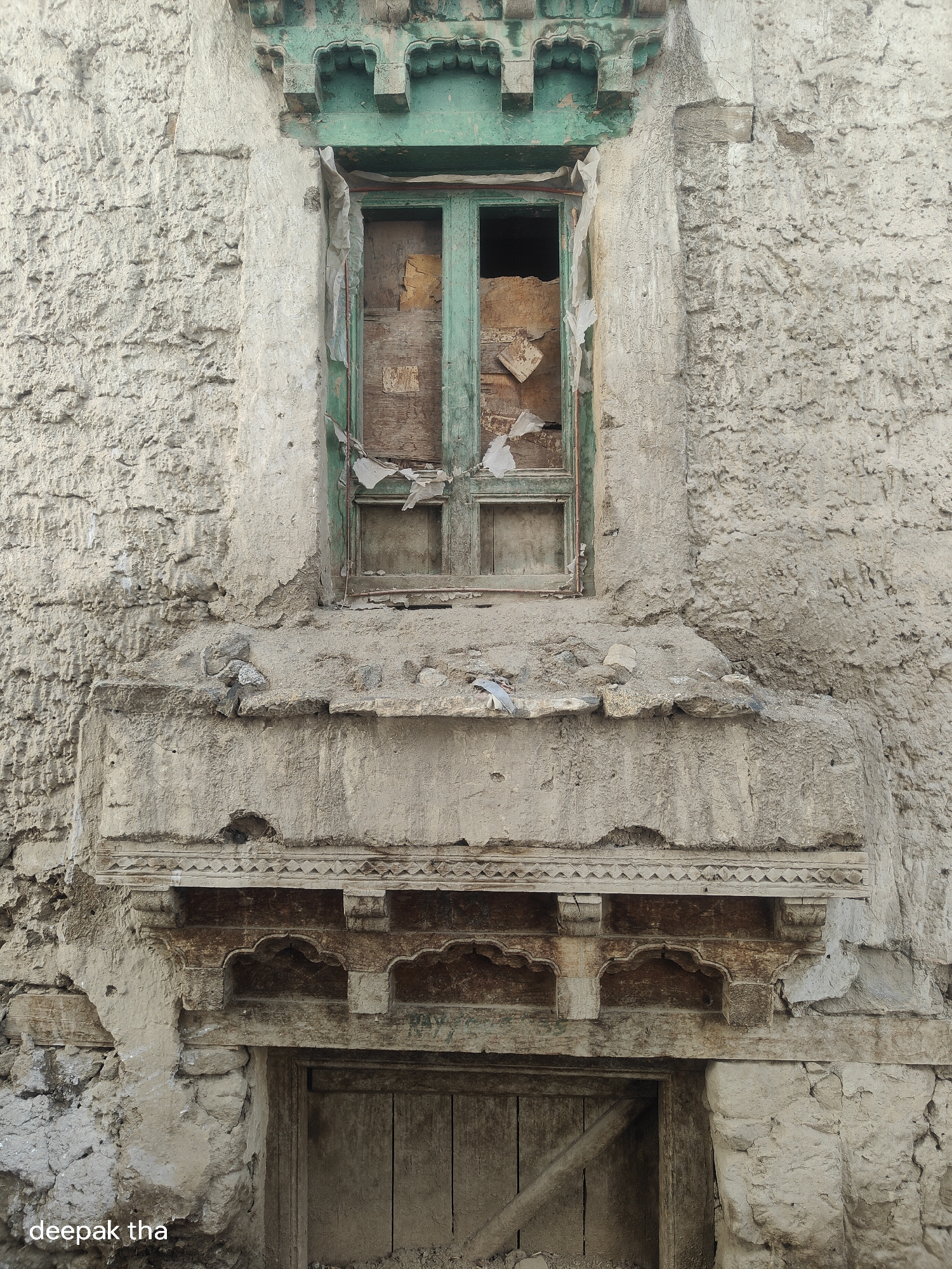 An exterior vertical shot captures a dilapidated, weathered window and doorway set into a rough, textured, light-colored stone or stucco wall. The window frame is painted a faded green and appears to be made of wood. The windowpanes are broken or missing, revealing the dark interior of the building and some debris, including pieces of wood and what looks like white plastic sheeting.
Below the window, there is a ledge or balcony structure, also made of stone or stucco, with a more ornate, carved wooden element beneath it. This wooden decoration features arched shapes and intricate patterns. Below this decorative element is a wooden doorway, with vertical planks and a diagonal wooden support beam. The entire structure appears aged and in a state of disrepair, with crumbling mortar, cracked surfaces, and peeling paint.
The scene is illuminated by natural light, casting subtle shadows and highlighting the textures of the materials. The bottom left corner of the image contains the watermark "deepak tha". The overall impression is one of abandonment or neglect of an old building in a harsh, possibly arid environment, which is consistent with the mountainous region of Leh, India.