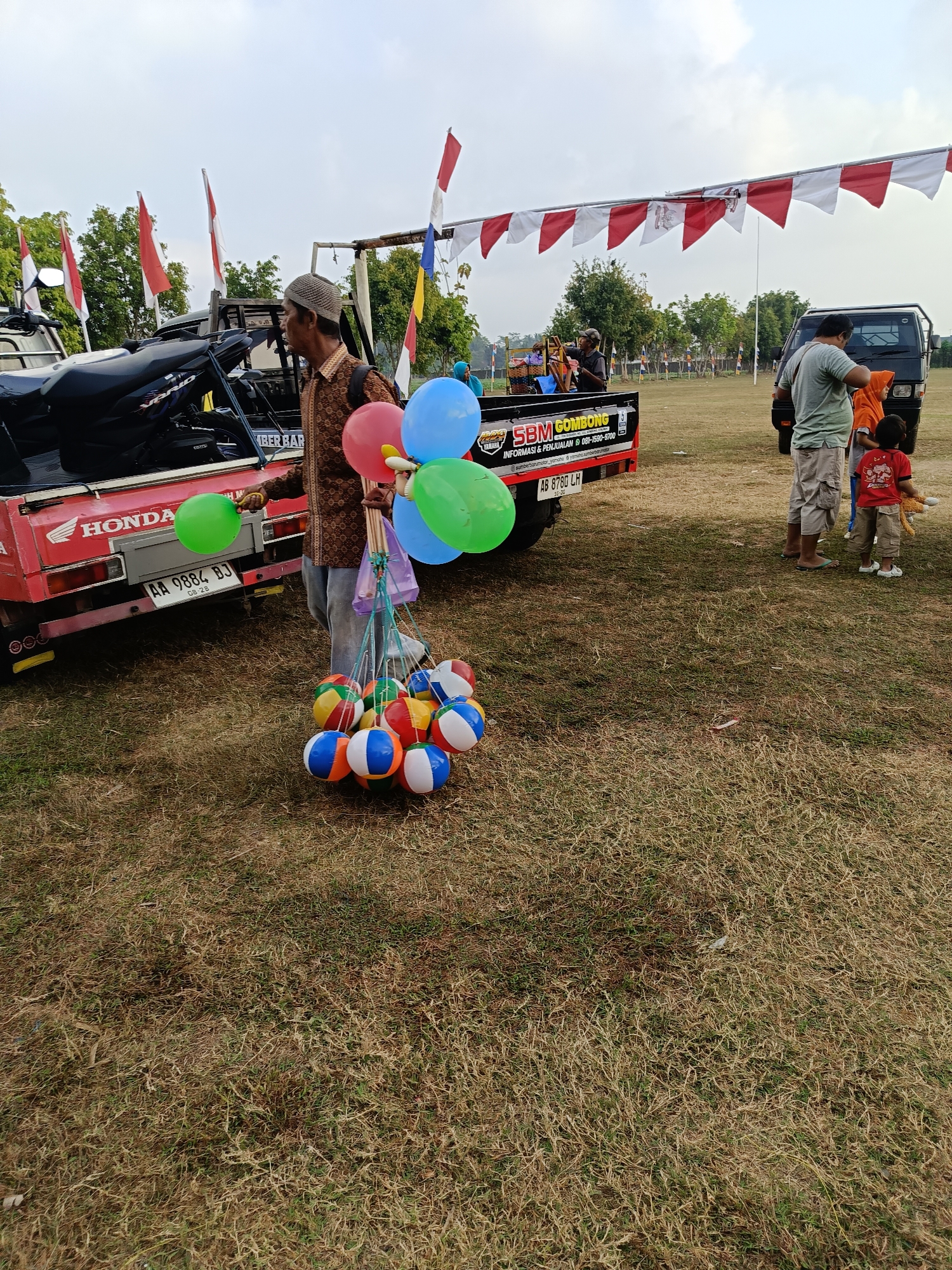 The image depicts a scene in Mangli, Indonesia, likely outdoors. In the foreground, a man wearing a patterned brown shirt and a koptan (a type of cap) is holding a bouquet of colorful balloons, primarily red, blue, and green. Below the balloons, he holds a cluster of striped inflatable balls, each featuring multiple colors like red, white, blue, and yellow. The man stands next to the back of a red pickup truck. The truck has a "HONDA" logo on its side and a license plate reading "AA 9884 BJ". A sign on the truck bed displays "SBM GOMBONG" and contact information.
In the mid-ground, several people are visible. To the right of the pickup truck, a man in a grey t-shirt and shorts stands with a child in a red t-shirt and beige shorts who is holding a stuffed toy. Behind them, a dark-colored van is parked. Further in the background, more people are scattered, and a line of red and white triangular flags is strung across poles, suggesting a festive or celebratory event. Red and white Indonesian flags are also visible, mounted on poles in the distance.
The environment is an open field covered in dry, brown grass. The sky is overcast with a muted, cloudy appearance. The overall impression is of a casual outdoor gathering or market, possibly in a rural or semi-rural area.