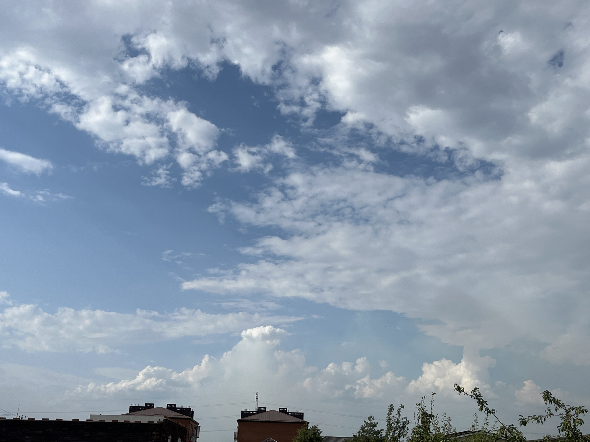 This is a photograph of a bright, partly cloudy sky over Taganrog City District, Russia. The sky is predominantly blue, with scattered cumulus clouds of varying sizes and densities. Some clouds are thin and wispy, while others are larger and more voluminous, with distinct white tops and darker undersides, suggesting potential for precipitation. In the foreground, the tops of several buildings are visible, including what appears to be a multi-story brick building with a dark roof and decorative elements along its facade. To the right, a few trees with green foliage are partially obscuring the view of the sky. Power lines are also visible stretching across the frame. The overall impression is that of a clear day with intermittent cloud cover.