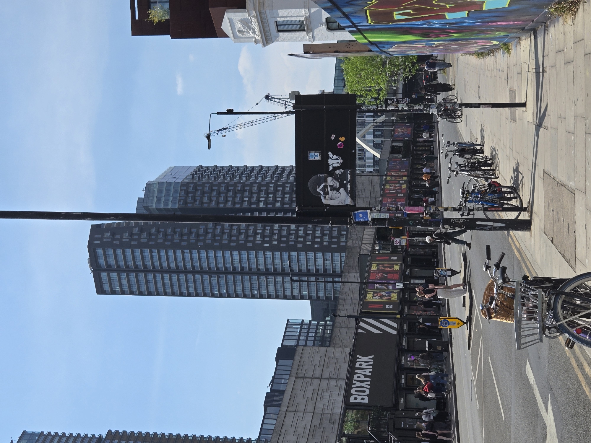 The image depicts a street scene in Shoreditch, London, United Kingdom, featuring Boxpark, a pop-up mall constructed of shipping containers. The foreground contains bicycles parked along the roadside. Pedestrians are visible walking along the sidewalk and in the road. A tall building and a construction crane are visible in the background. Graffiti adorns the wall along the sidewalk on the right side of the image. Advertisements and posters are attached to the structure of Boxpark.