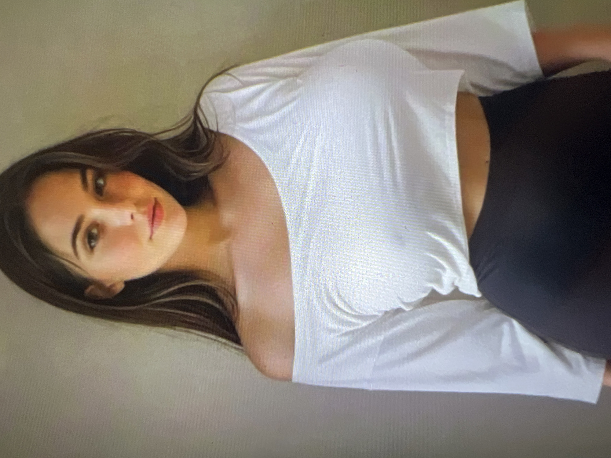 A young woman with long, dark hair is shown from the chest up, wearing an off-the-shoulder white top and a black bottom. She is standing in front of a plain, light-colored wall. Her head is slightly tilted, and she is looking directly at the camera.