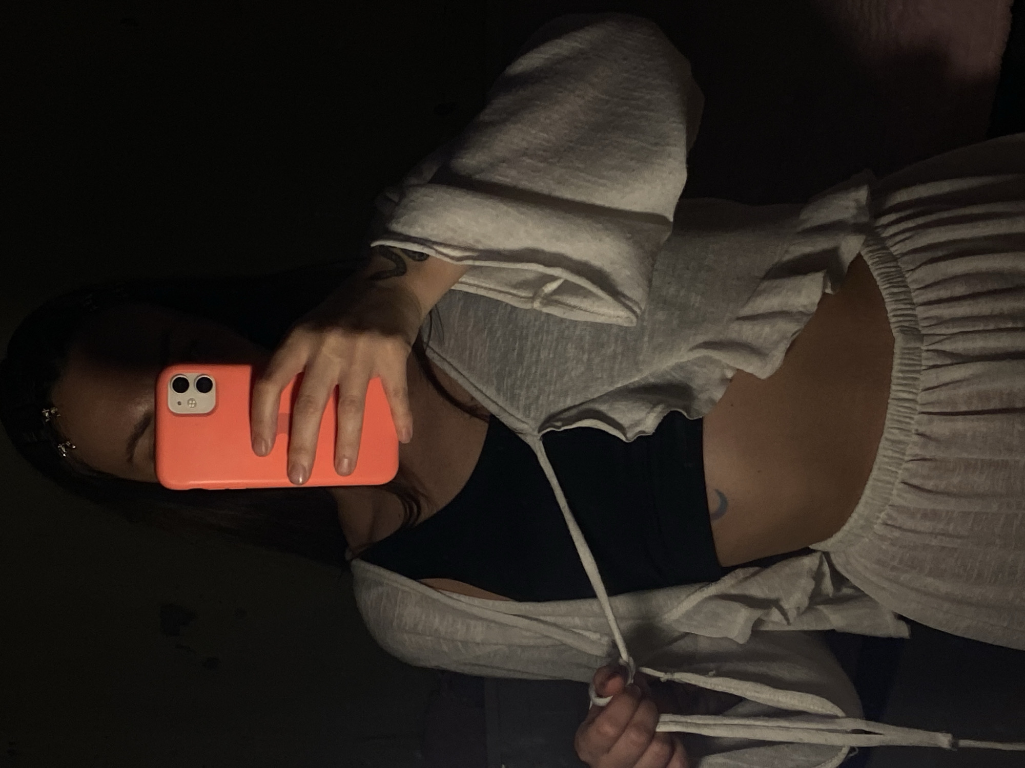 A young woman is depicted in a dimly lit indoor setting in Votkinsk, Russia, captured in a self-portrait or mirror selfie. She holds a bright coral/orange smartphone with her right hand, positioned to obscure most of her face. The phone prominently displays two camera lenses. Her dark hair is visible, styled with small metallic accessories near her temple.
She is dressed in a casual outfit consisting of a light grey, loose-fitting, long-sleeved top that is open at the front and secured by white ties at the waist. Beneath this, she wears a black bra or crop top, revealing her midriff. Her lower attire includes light grey pants featuring a gathered, elastic waistband. Distinctive features include a crescent moon tattoo on her right side, just above the hip, and another darker tattoo on her left forearm, visible near the sleeve cuff.
The low lighting suggests the image was taken during the evening or nighttime. The background is largely dark and indistinct, offering no specific architectural or environmental cues for Votkinsk, Russia, beyond a generic indoor space. No visible text is present in the image.