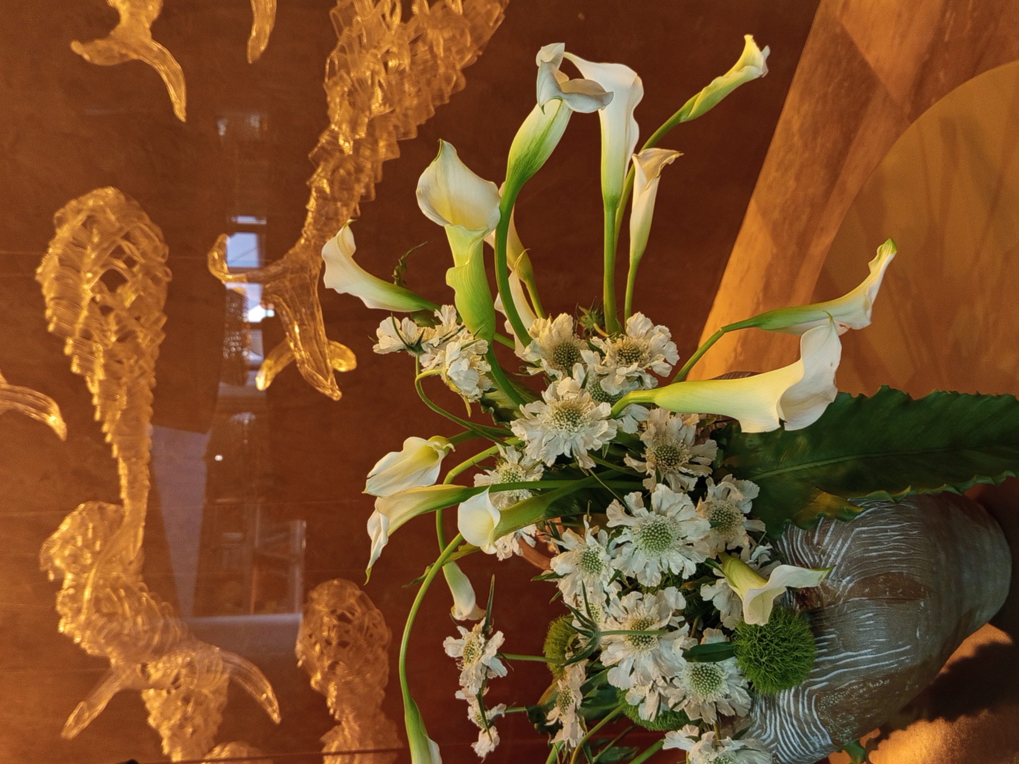 The image displays a floral arrangement in a textured vase. The bouquet features white calla lilies and smaller white flowers, set against a dark, reflective background. The background is adorned with abstract, translucent decorative elements that appear to be made of glass or resin, resembling stylized clouds or aquatic forms. The overall lighting is warm and ambient, casting soft shadows. There are no people visible in the image. The scene is indoors, likely in a setting with a modern or artistic interior design, possibly a hotel lobby, gallery, or a well-appointed residence. Without specific visual cues to landmarks or recognizable architecture, it is impossible to determine if the image was taken in Saint Petersburg, Russia, or any other specific city.