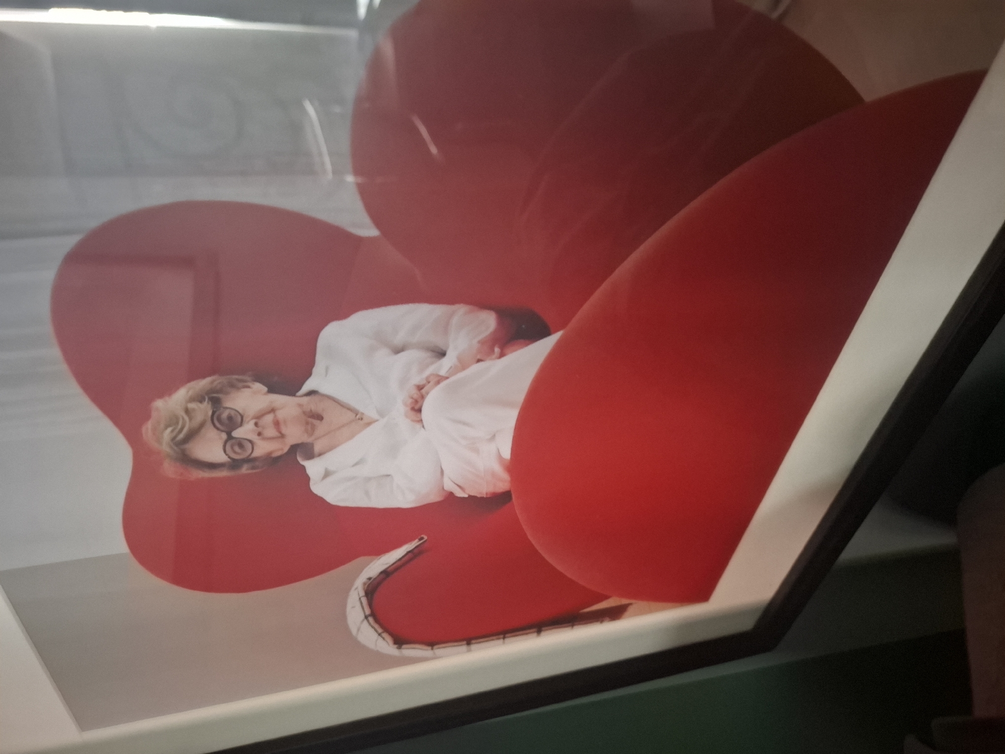 A framed photographic portrait depicts an elderly woman seated in a large, red, heart-shaped chair. The woman has short, blonde hair, wears round glasses, and is dressed in white. Her hands are clasped in her lap. The chair appears to be made of a smooth, possibly upholstered material. The background is out of focus, with hints of a room that includes a wall and possibly a window or light source. The photograph is displayed in a gallery or personal space, as indicated by the frame and the surrounding environment, which shows elements of interior decor. The image appears to be located in Vincennes, France.