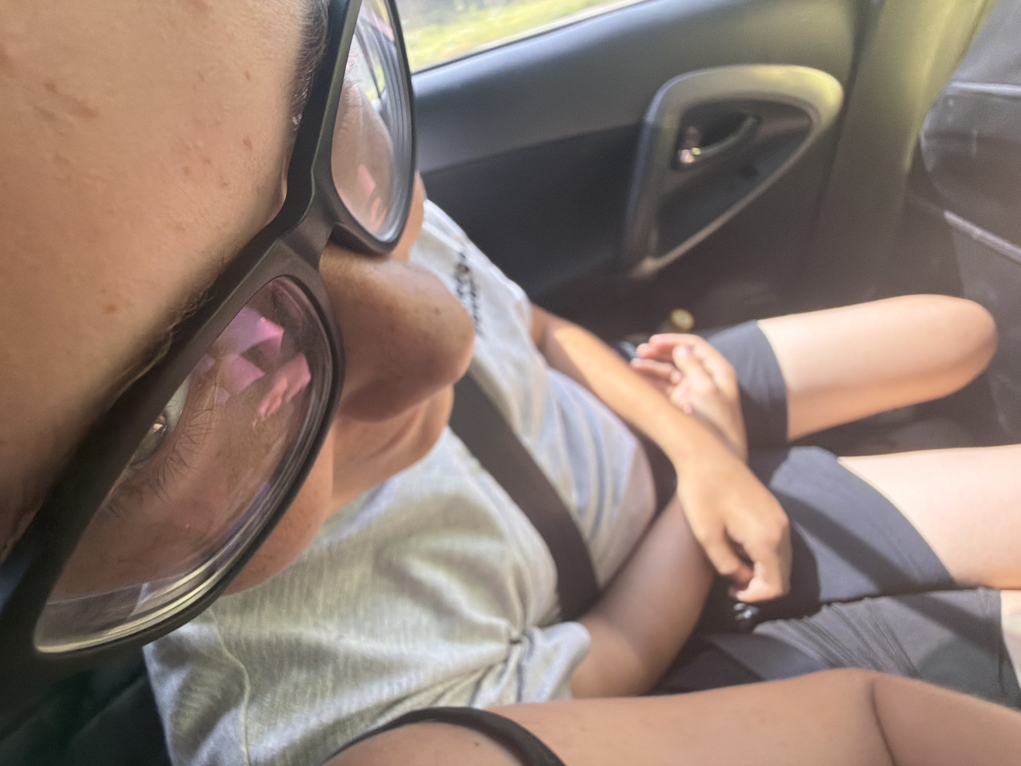 The image shows a close-up, angled shot from inside a car, looking down at a person wearing glasses and a grey t-shirt. The person's hands are clasped in their lap, and they are wearing dark shorts. The car interior is visible, including a black door panel and a seatbelt. The lighting suggests it is daytime, with sunlight reflecting off the glasses. The background is blurred, but appears to be foliage, indicating the car is likely in a rural or suburban area. The location of Kruglovka, Russia, is not determinable from the image.