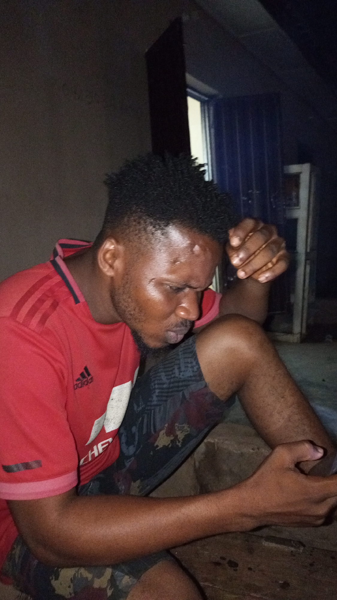 A man sits in dim lighting. He wears a red t-shirt with a white logo and grey patterned shorts. He is holding something in his right hand and his left hand is clenched near his face. The location is likely in Sabon Tasha, Nigeria, based on context provided (not visible in the image itself). There is a doorway or window behind him. The background is dark and indistinct.