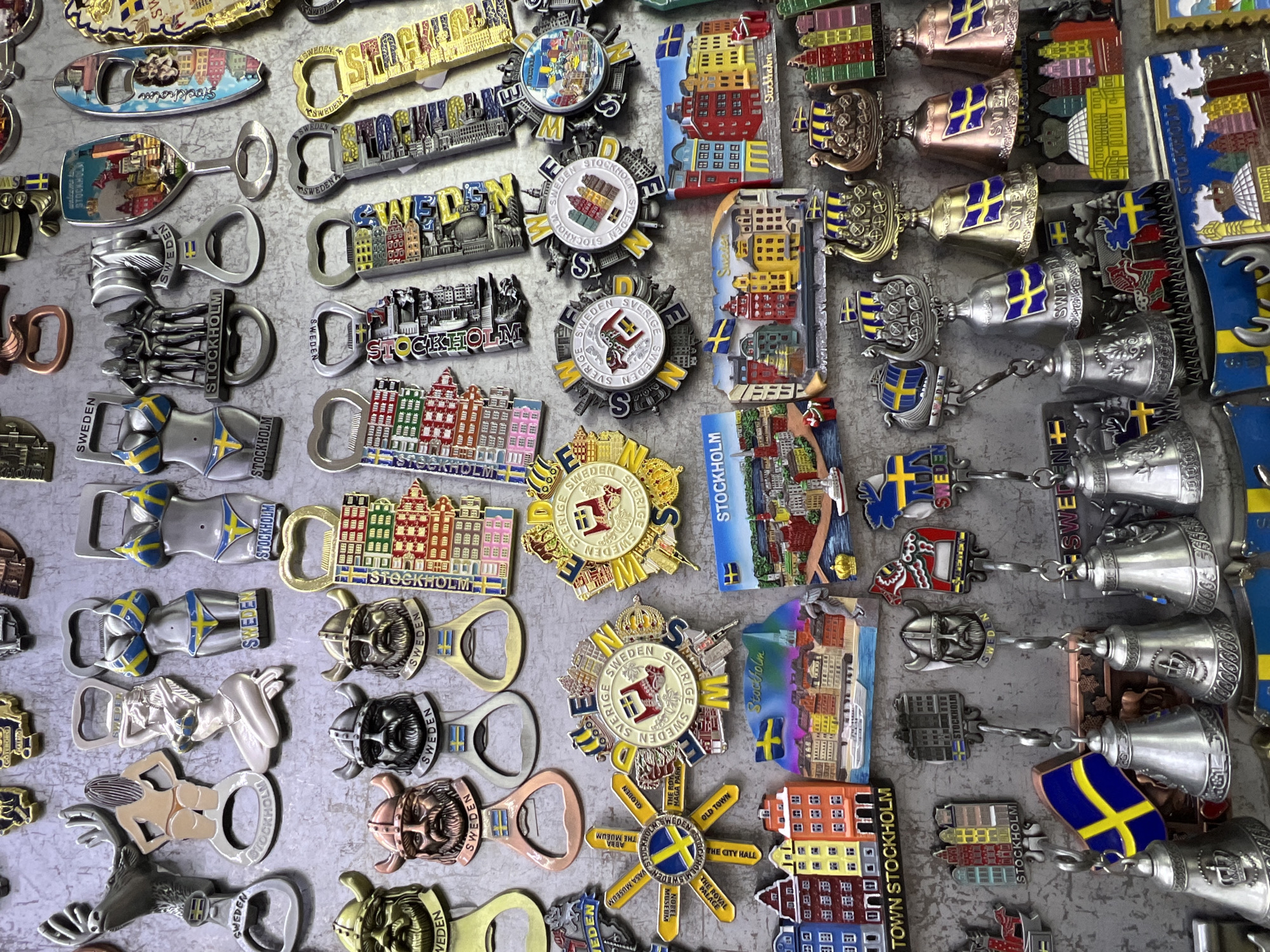 This image displays a collection of Swedish souvenir magnets and bottle openers arranged on a textured gray surface. The items are predominantly themed around Stockholm and feature iconic Swedish imagery such as the Swedish flag, Viking motifs, and depictions of Stockholm's architecture. There are no people or actions depicted. The collection includes bottle openers shaped like women in Swedish national swimwear, Viking heads, and miniature buildings, as well as magnets featuring panoramic views of Stockholm, its landmarks, and various decorative designs. The overall impression is that of a display of tourist merchandise.