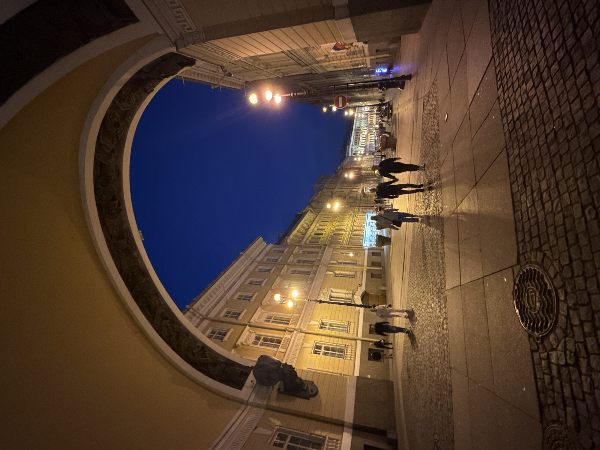 The image captures an evening view looking down a cobblestone street in Saint Petersburg, Russia, framed by a large architectural archway. The sky is a deep twilight blue. On either side of the street are buildings with illuminated windows, displaying a neo-classical architectural style with ornate detailing. Streetlights, some with multiple lamps, cast warm light onto the pavement.
Several individuals are walking along the street. In the foreground, a couple holds hands as they walk away from the camera. Further down the street, other pedestrians can be seen. Decorative planters and lampposts line the sidewalks.
The foreground features a circular metal manhole cover with a decorative pattern. The ground is paved with cobblestones and large rectangular stone slabs. The overall scene depicts a lively urban environment transitioning from day to night. The architectural style of the buildings and the presence of the archway strongly suggest a location within the historical center of Saint Petersburg, possibly near a palace or a grand public building.