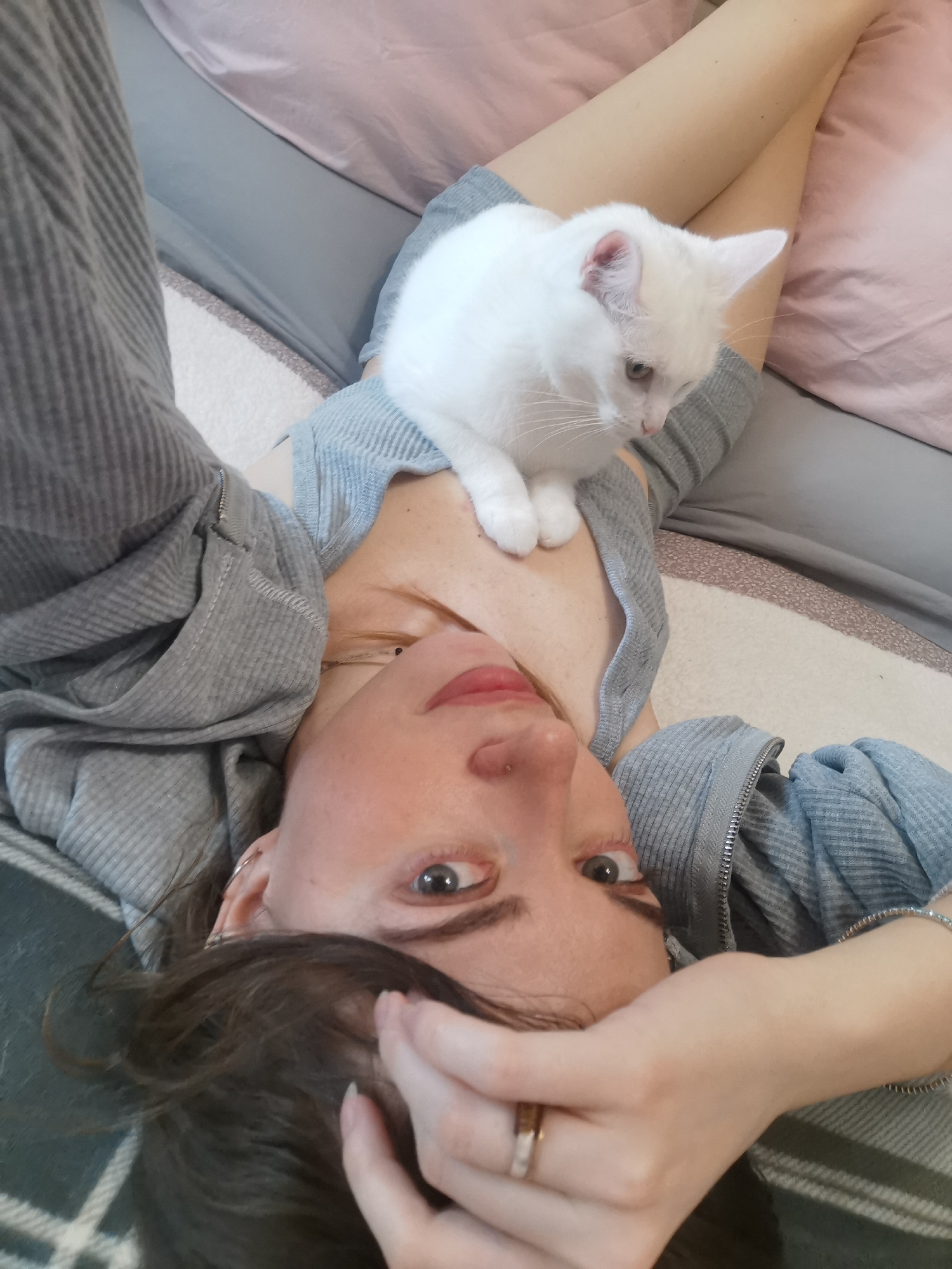 A woman is lying on her back, looking up at the camera. Her right hand is near her forehead, and she is wearing a ring on her ring finger. A white cat is sitting on her lap. She is wearing a gray sweater. The background includes a gray pillow, a pink sheet, and a gray and green blanket. The location is unknown.