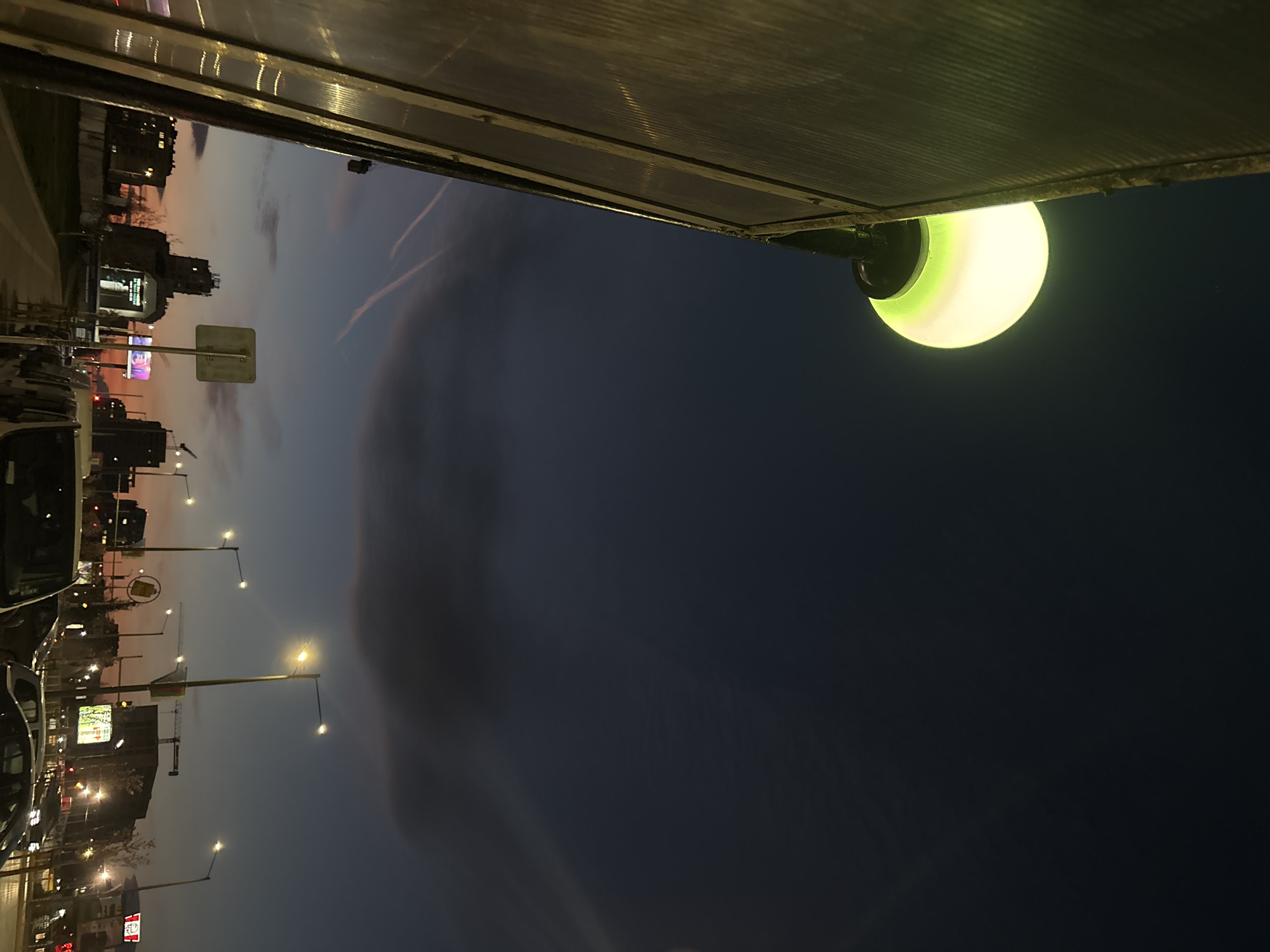 The image depicts a nighttime urban scene in Sofia, Bulgaria. A row of streetlights illuminates a street lined with buildings, including a KFC. The sky is dark, with streaks of clouds near the horizon. A street lamp with a globe fixture is prominently positioned in the foreground. A metallic structure, possibly a roof or awning, frames the upper portion of the view. Cars are parked along the left side of the street.