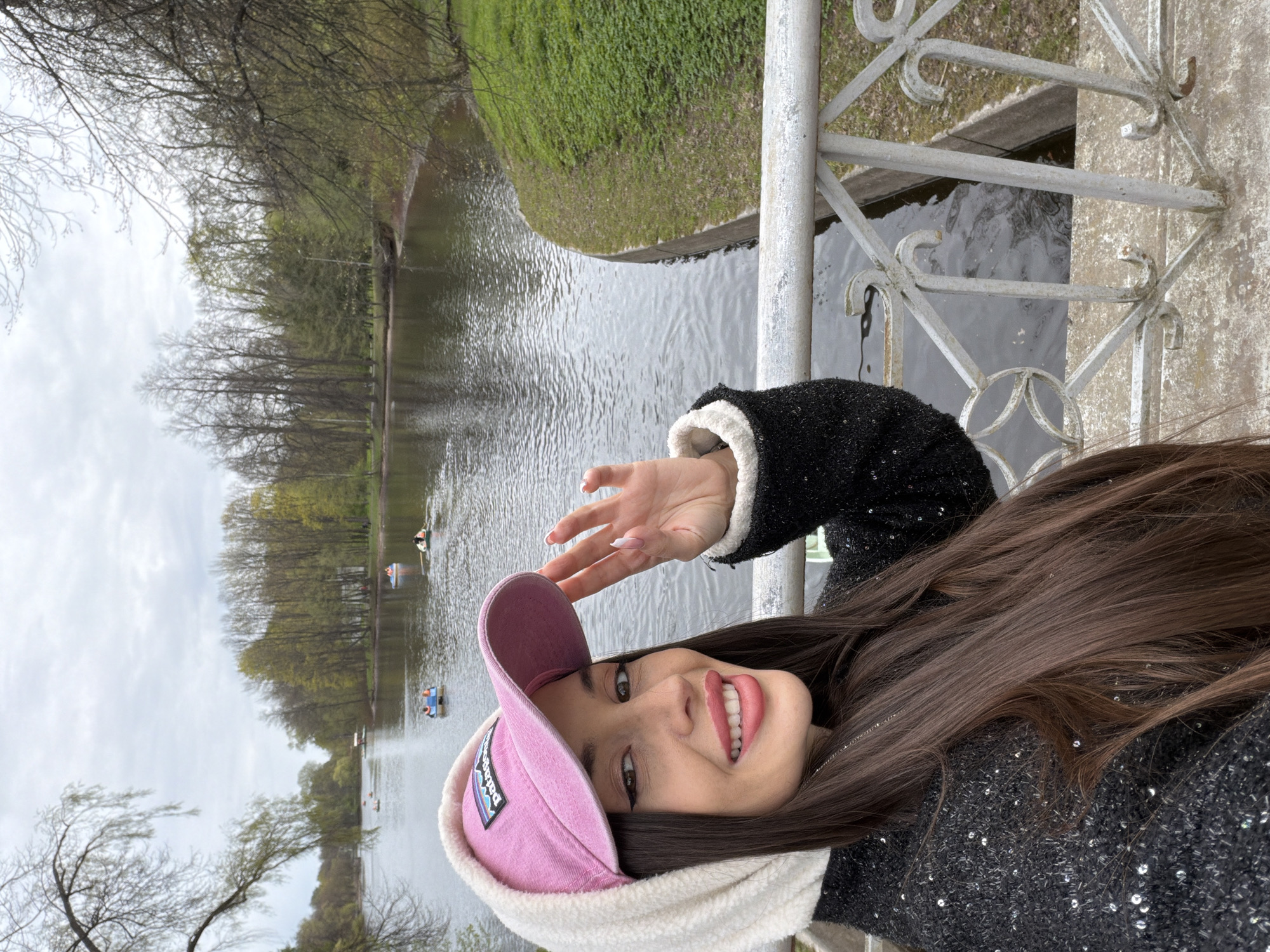 A young woman poses for a selfie on a bridge in Saint Petersburg, Russia. She wears a pink cap with a white fleece lining and a dark, sequined jacket with white trim. Her long, brown hair is visible. The background features a body of water, possibly a lake or river, with trees lining the far bank. Several small boats with people in them are visible on the water. A metal railing of the bridge is partially visible on the right side of the frame.