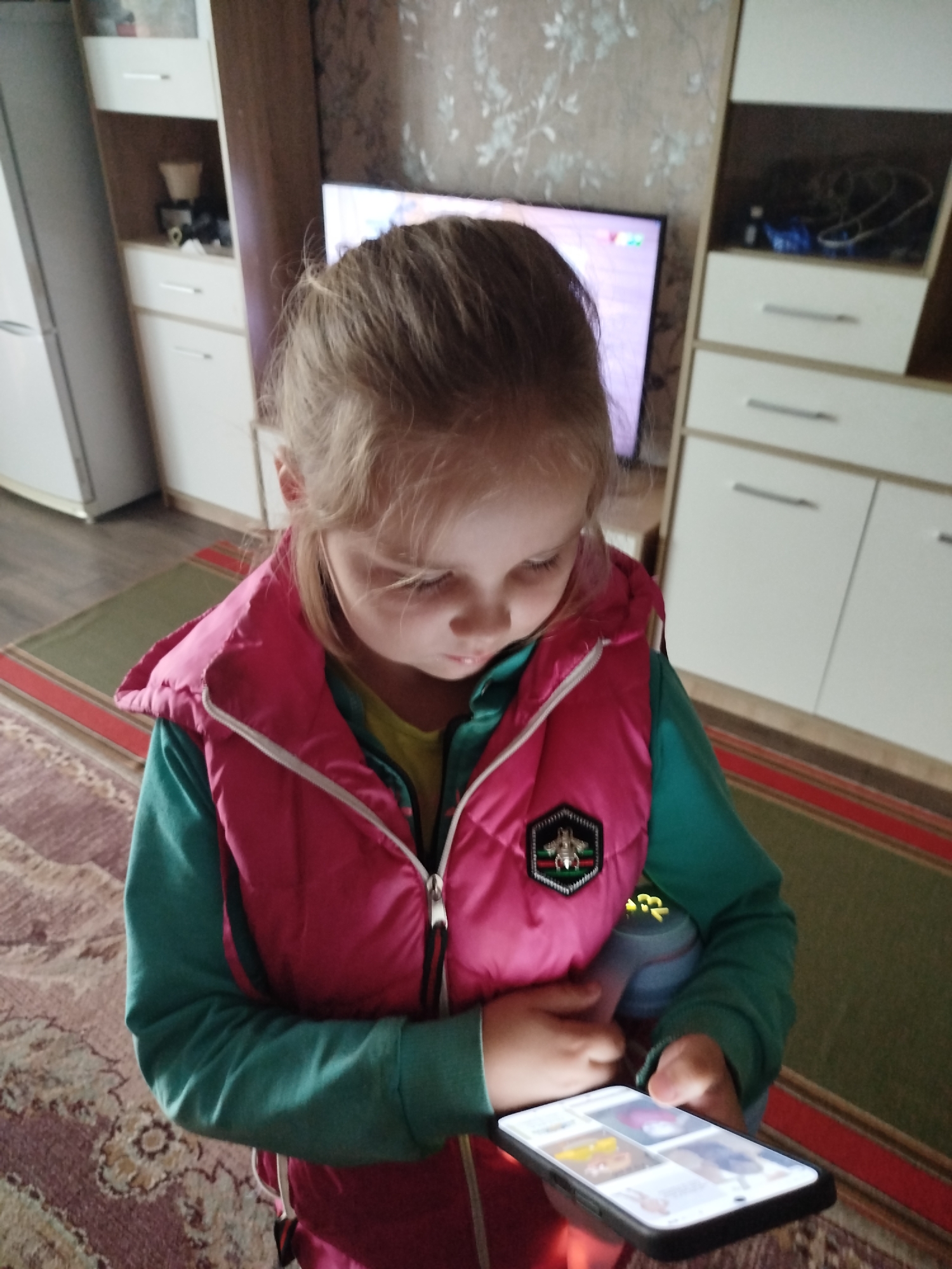 A young girl with blonde hair, wearing a pink vest over a green long-sleeved shirt, is looking down at a smartphone in her hands. She is standing in what appears to be a living room, with a television in the background and furniture, including a tall cabinet, visible. A rug is on the floor. The image is taken from a slightly elevated angle. The girl's expression is focused on the screen. The environment suggests a domestic setting within the Крывасельскі сельскі Савет, Belarus.