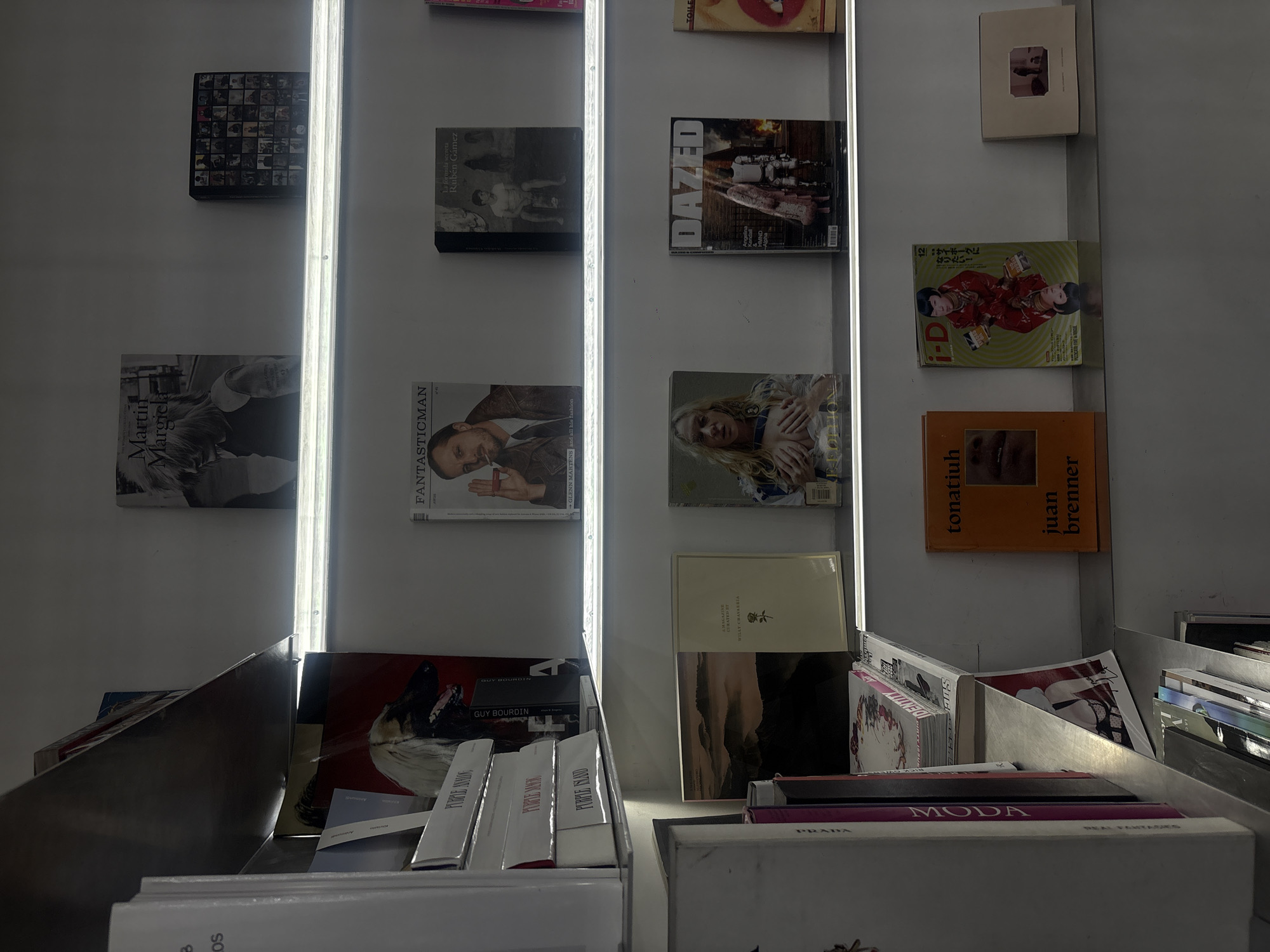 This image, taken in Mexico City, Mexico, shows a retail setting with vertically arranged books and magazines on white shelves. The books, including titles like "Martin Margiela," "FANTASTICMAN," "DAZED," and "tonatiuh juan brenner," are positioned on the shelves, some in stacks and others individually. Beneath the shelves are containers holding more books and magazines, including titles such as "PURPLE ISLAND," "PURPLE AMONG," "MODA," and "PRADA." The environment is illuminated by vertical light bars, which create bright reflections. The photo captures the atmosphere of a bookstore or retail space focused on design and fashion publications.