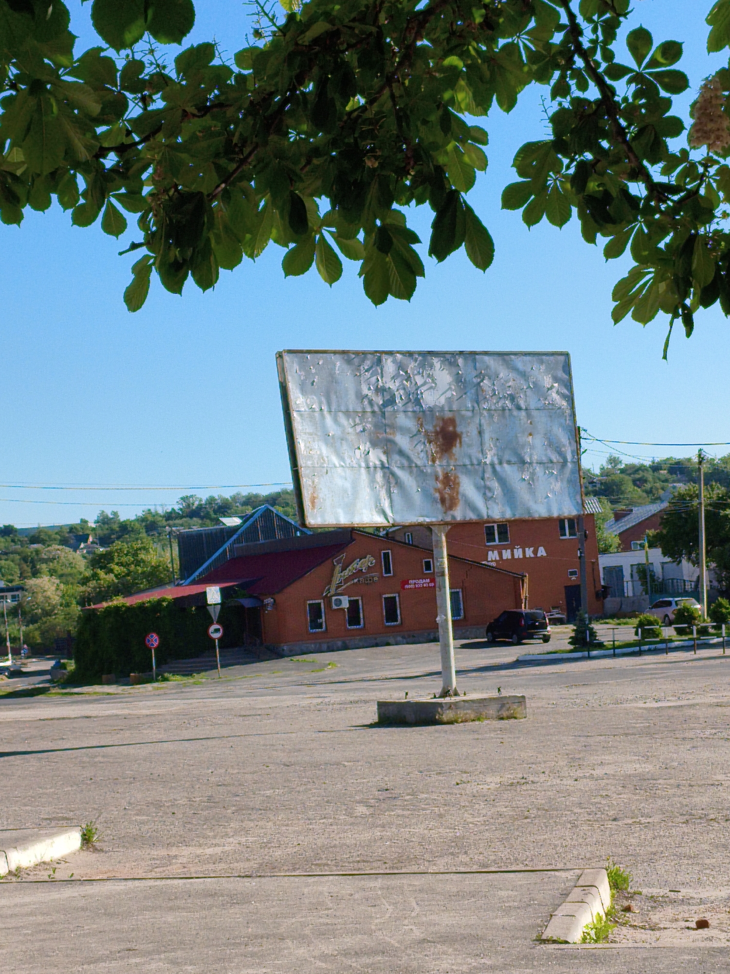 The image captures a roadside scene in Velyka Bahachka, Ukraine, under a clear, bright blue sky, suggesting daytime, likely morning or early afternoon. The foreground is dominated by a large, empty asphalt parking lot or open space, with a concrete curb in the bottom right corner. Overhanging green leaves from a tree, possibly a chestnut, frame the top of the photograph.
A prominent feature in the midground is a large, rectangular billboard made of corrugated metal, showing considerable weathering, rust, and peeling paint. It stands on a single concrete pillar in the center of the frame, currently advertising nothing. Behind the billboard are two brick buildings with red roofs. The building on the left, which appears to be a cafe or shop, has a sign displaying "Джерело" (Djerelo) and smaller text below that reads "ПРОДАМ" (FOR SALE), a phone number "(050) 933 93 69", and "ПРОДАЖ" (SALE). The building to its right has the word "МИЙКА" (CAR WASH) prominently displayed on its facade. A dark-colored car is parked between these two buildings.
Several small road signs are visible near the left building: a red circular sign with a white horizontal bar (indicating 'no entry' or 'forbidden') and another circular sign with a red border and a '0' inside, possibly indicating a parking restriction or local regulation. The background features a hilly landscape covered with lush green trees and scattered houses, indicating a semi-rural setting. The overall impression is one of a functional, albeit slightly rundown, commercial area in a small town.