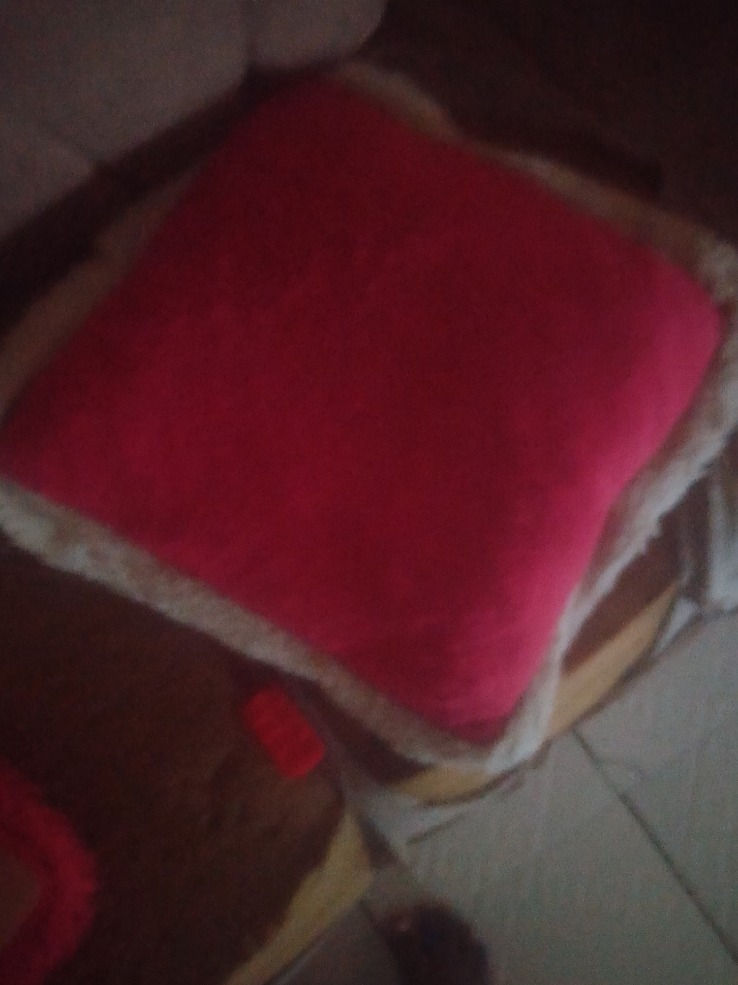 The image shows a close-up of a plush, square pillow with a bright pink front and a cream-colored fluffy trim. The pillow appears to be resting on a darker surface, possibly a rug or bedding, with a brown and beige striped pattern visible underneath. The lighting suggests it is indoors, and given the context of Jauro Yinu, Nigeria, it's likely a domestic setting. There is a small red object, possibly another toy or accessory, to the left of the pillow. The overall impression is one of comfort and playfulness, perhaps a child's room. No people are visible, nor is there any text. The image is slightly blurry, giving it a soft, dreamy quality.