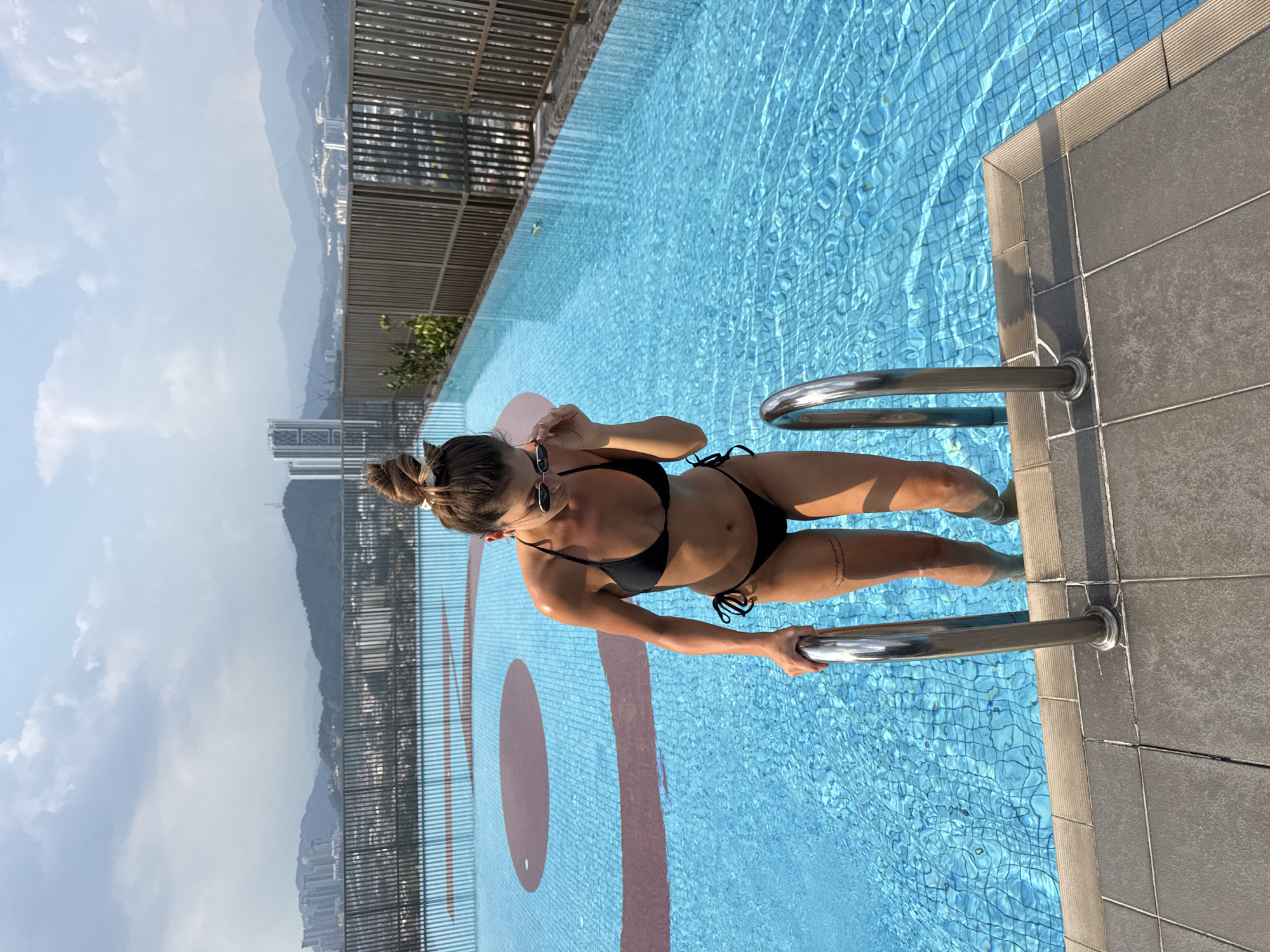 A woman in a black bikini is standing in a swimming pool, holding onto a metal ladder. She is wearing sunglasses and has her hair up in a bun. A pink inflatable ring is behind her. The pool is tiled with a light blue mosaic and has a large red oval design on the bottom. The pool is outdoors, with a view of buildings and mountains in the background. The location appears to be in Kuala Lumpur, Malaysia.