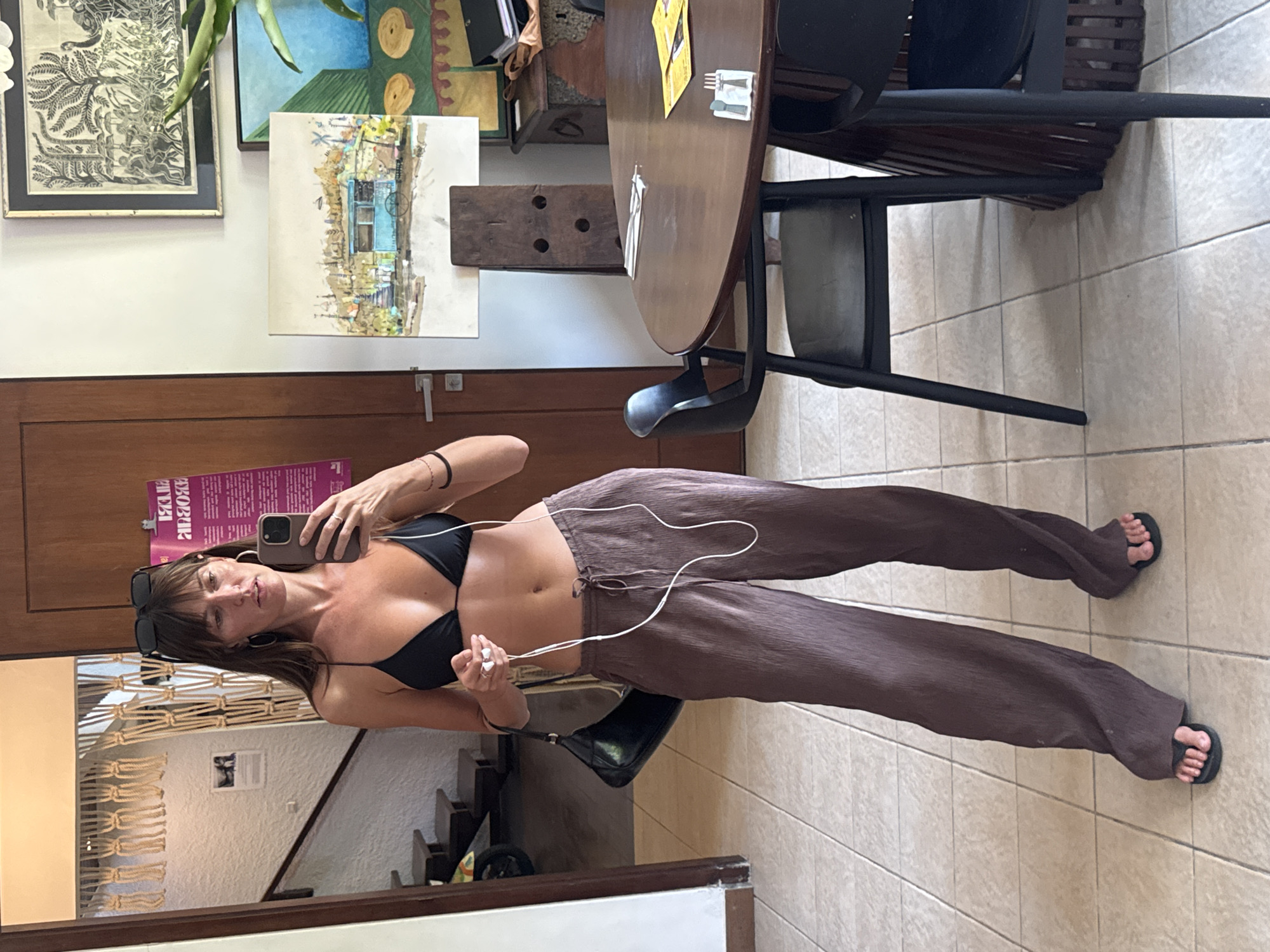 A woman is taking a mirror selfie in a room. She is wearing a black bikini top and brown wide-leg pants. She has on black flip-flops and is holding white earbuds in her hand. A black purse is slung over her left shoulder. She is wearing large hoop earrings and sunglasses are on top of her head. The room appears to be an indoor common area, possibly a cafe or restaurant, with tiled floors and a wooden door. There is a round wooden table with chairs in the background, along with several pieces of art on the wall. A macrame wall hanging is visible in the background reflected in the mirror. The image was taken in Mengwi, Indonesia.