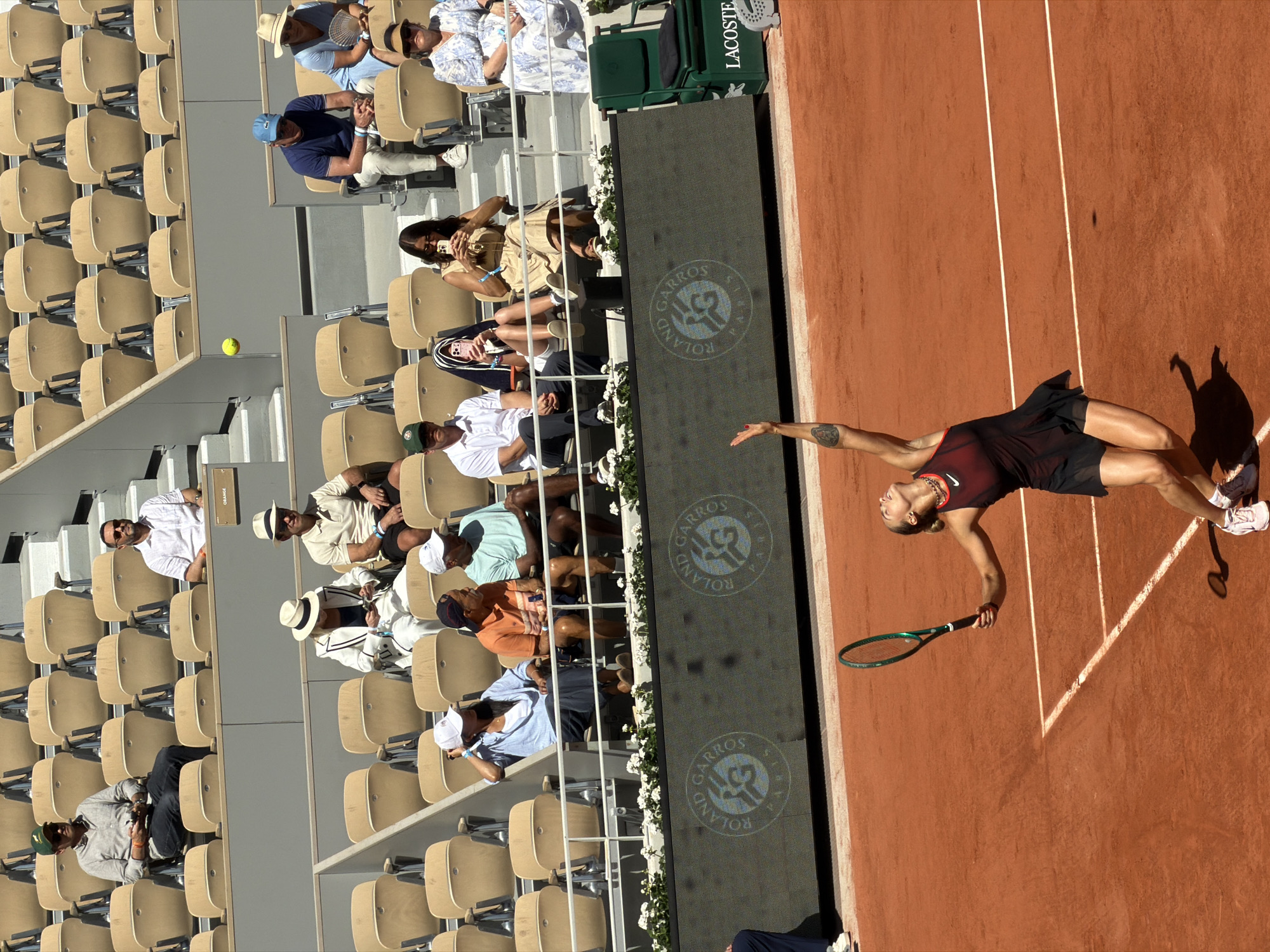 The image shows a tennis match at the Roland Garros French Open in Paris, France. A tennis player, wearing a dark red top and black shorts, is in the middle of serving. The clay court is reddish-brown, with white lines marking the boundaries. Behind the court are rows of seats filled with spectators. A green "Lacoste" sign is partially visible at the top of the frame. The crowd is diverse, with people wearing various outfits, some in hats. A tennis ball is visible in the background.